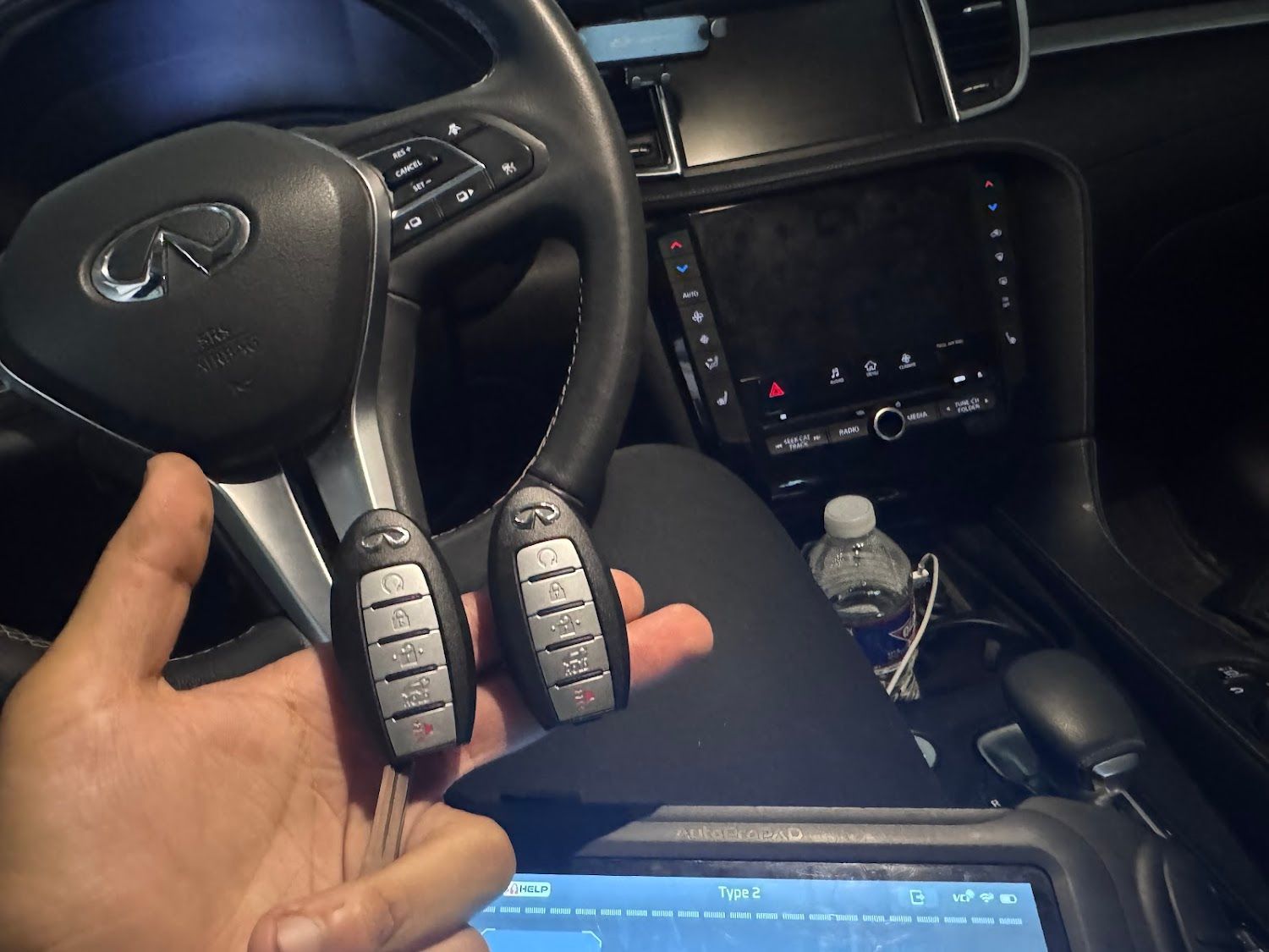 Hand holding two Infiniti car keys inside a vehicle's interior near the dashboard.