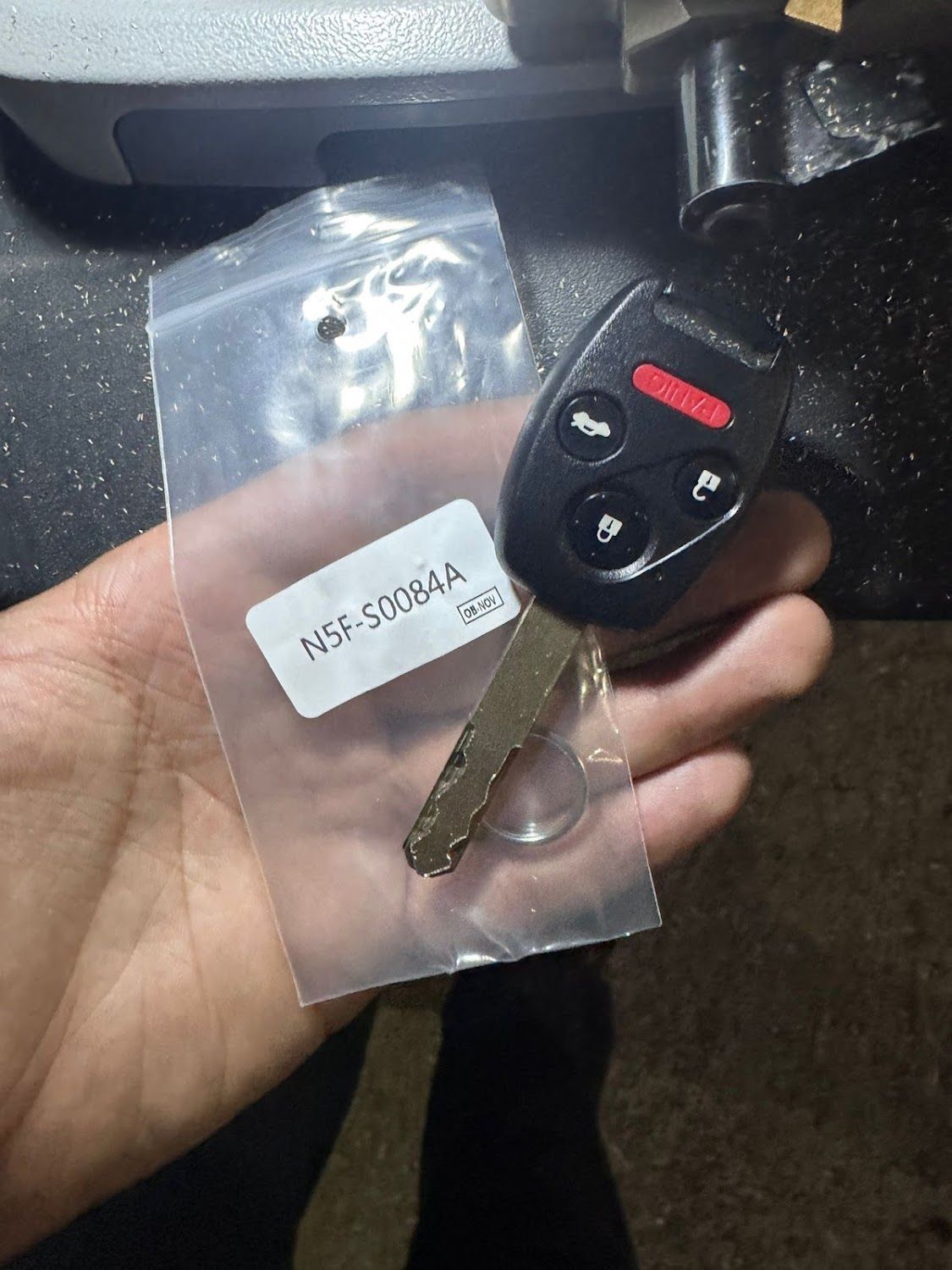 Hand holding a new car key and remote in a plastic bag with a label, near a machine.