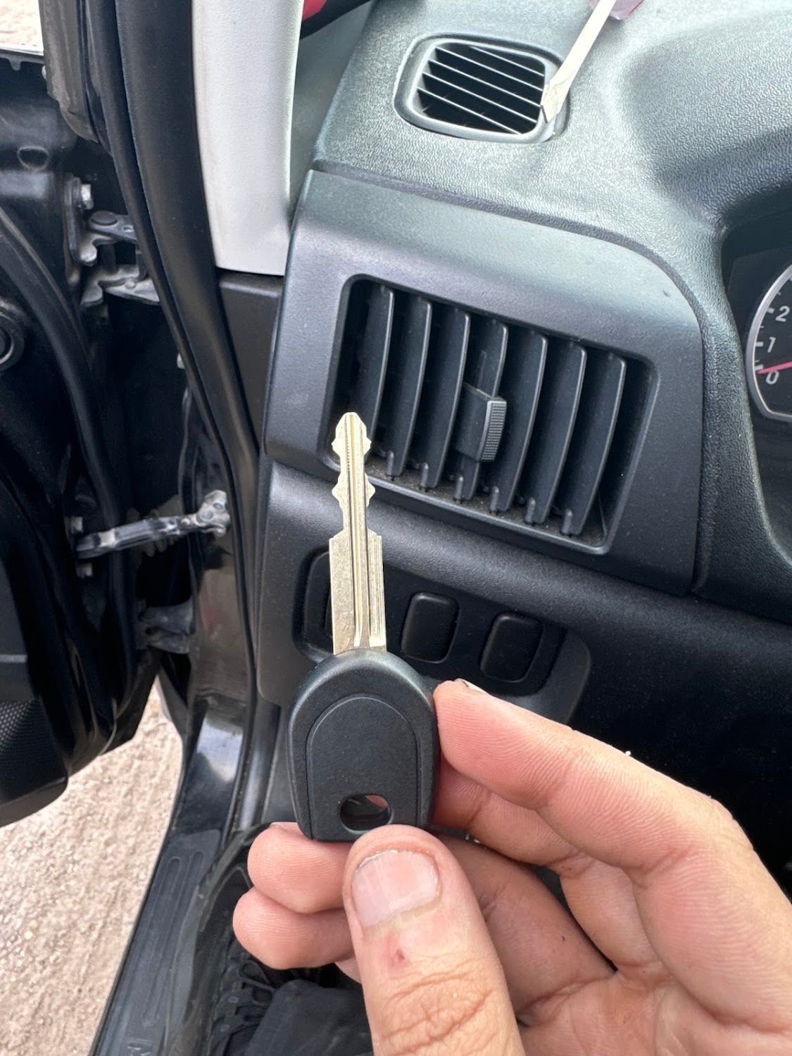 Hand holding a car key near a dashboard vent. Black dashboard and doorframe are visible.