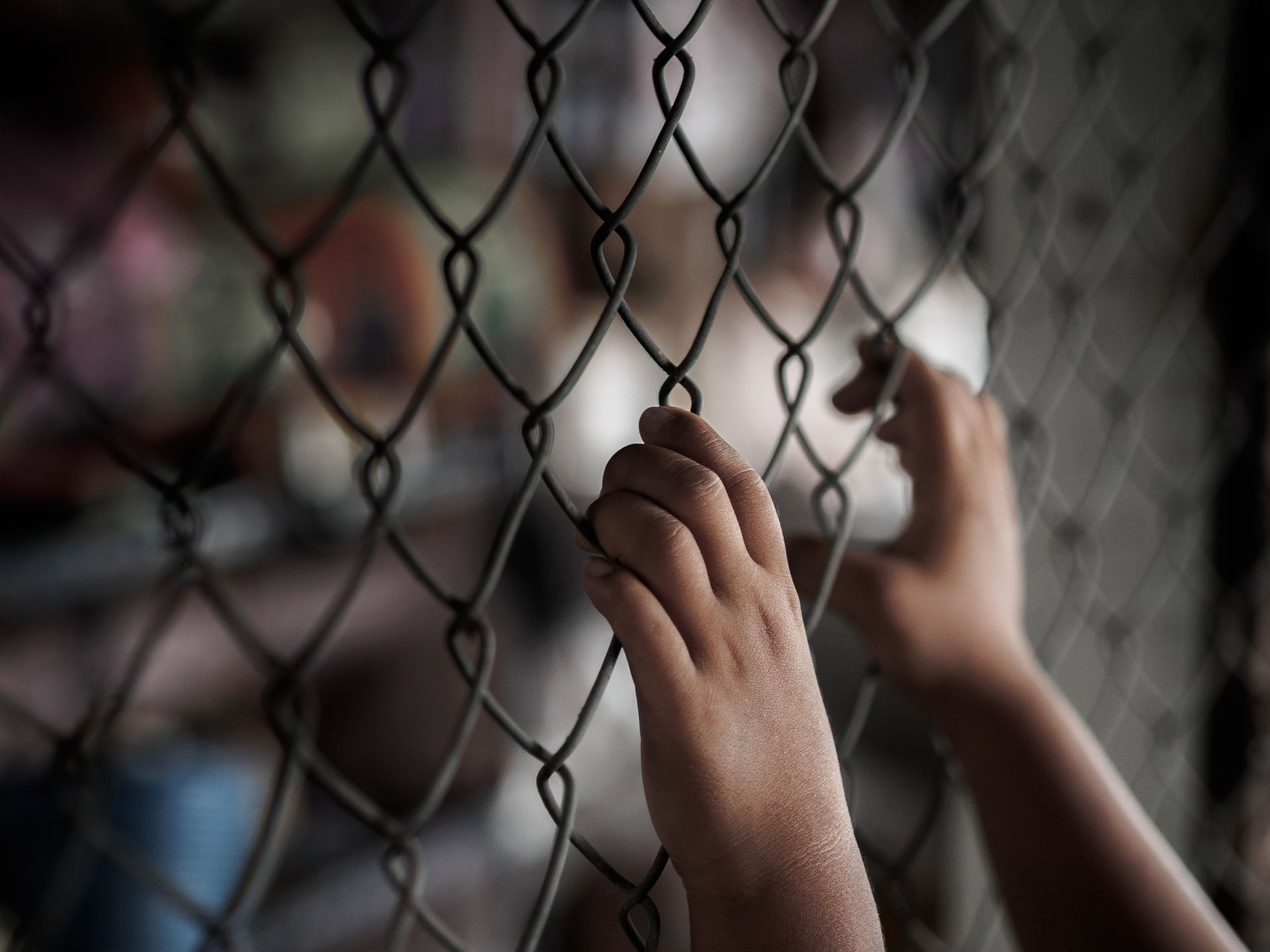 Child's hands grasping chain-link fence. Blurred background suggests detention setting.