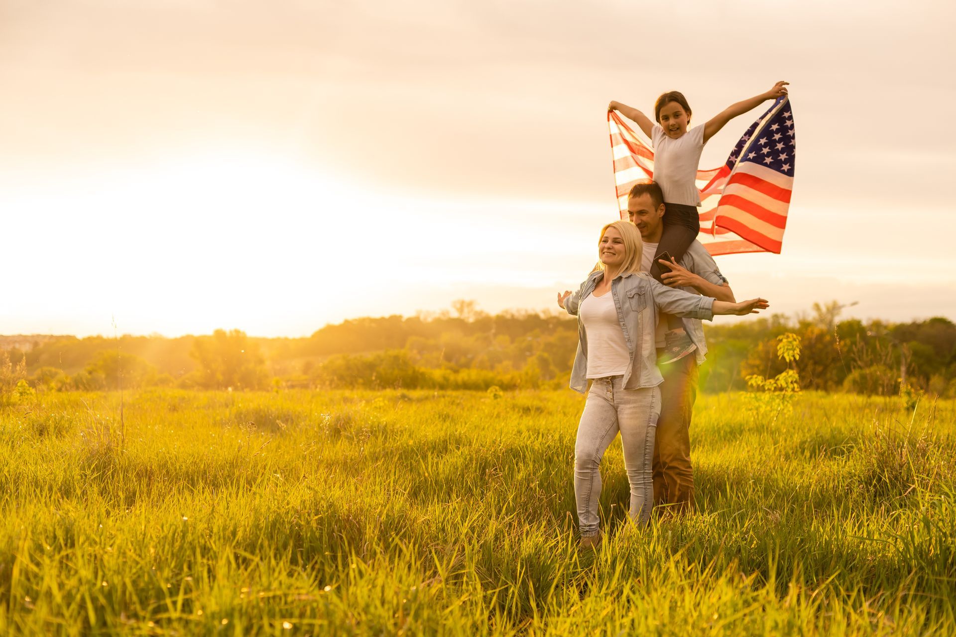 Family with child on shoulders holding US flag in a sunny field.