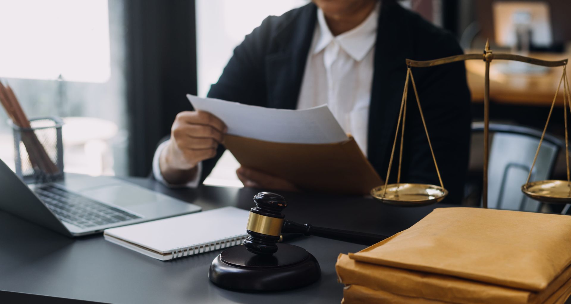 A person in business attire holds legal documents at a desk featuring a gavel, a laptop, and a scale of justice.