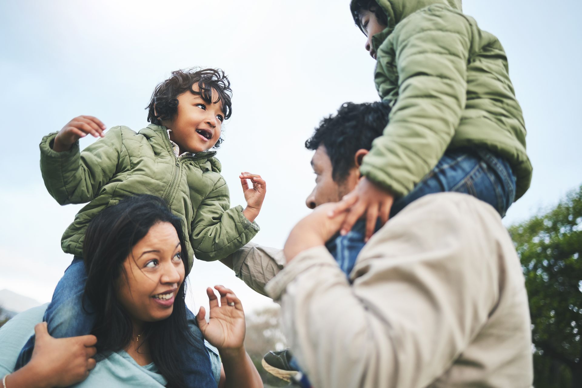 Family outdoors, two children on parents' shoulders, smiling, wearing jackets.