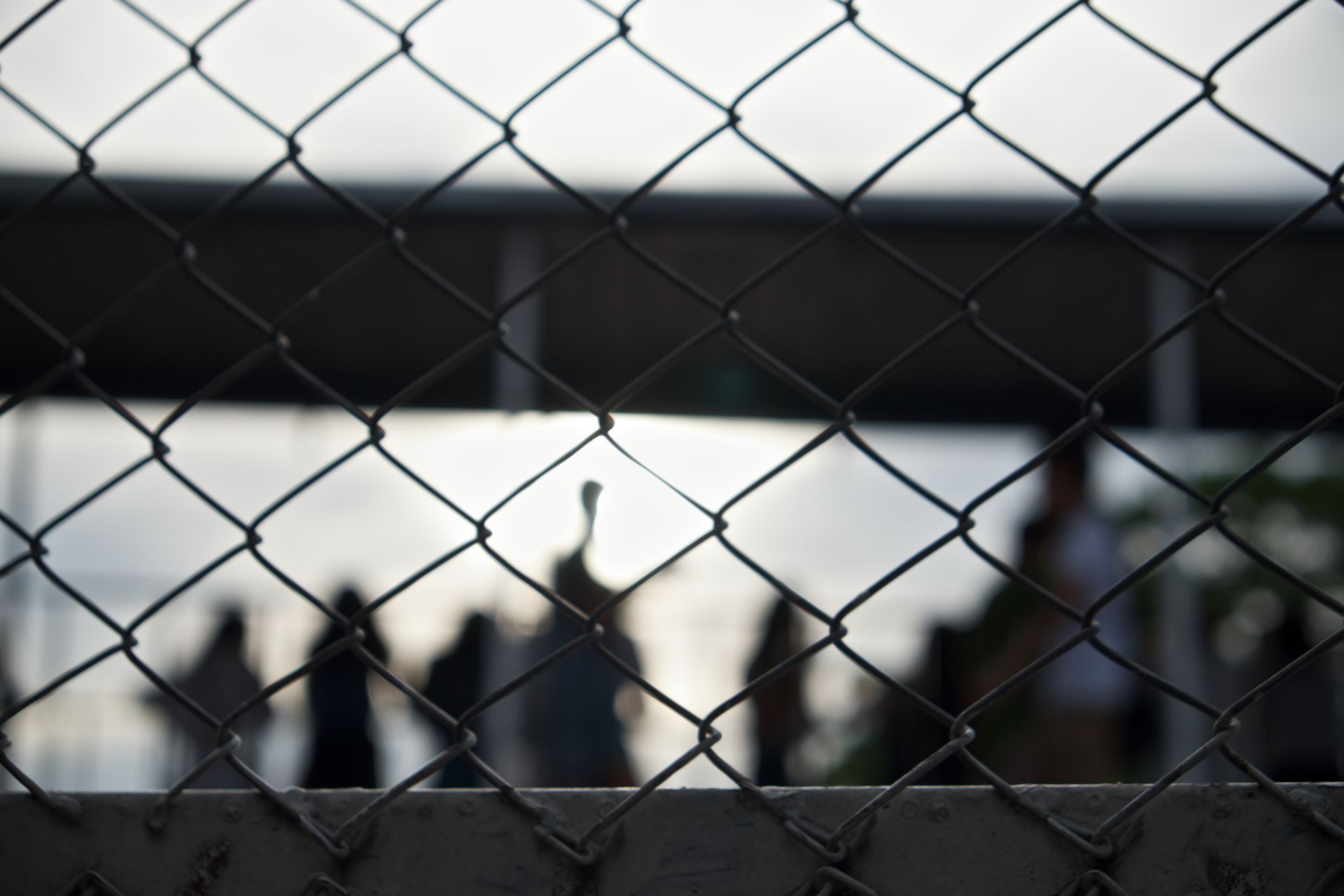 Close-up of chain-link fence, with blurred figures visible in the background against a bright sky.