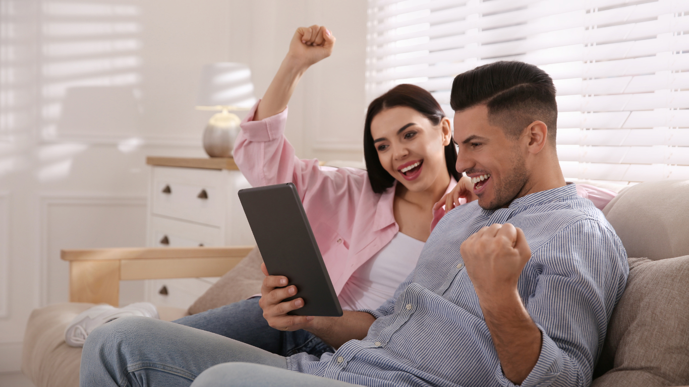 Smiling couple on a couch, cheering as they look at a tablet in a bright living room.