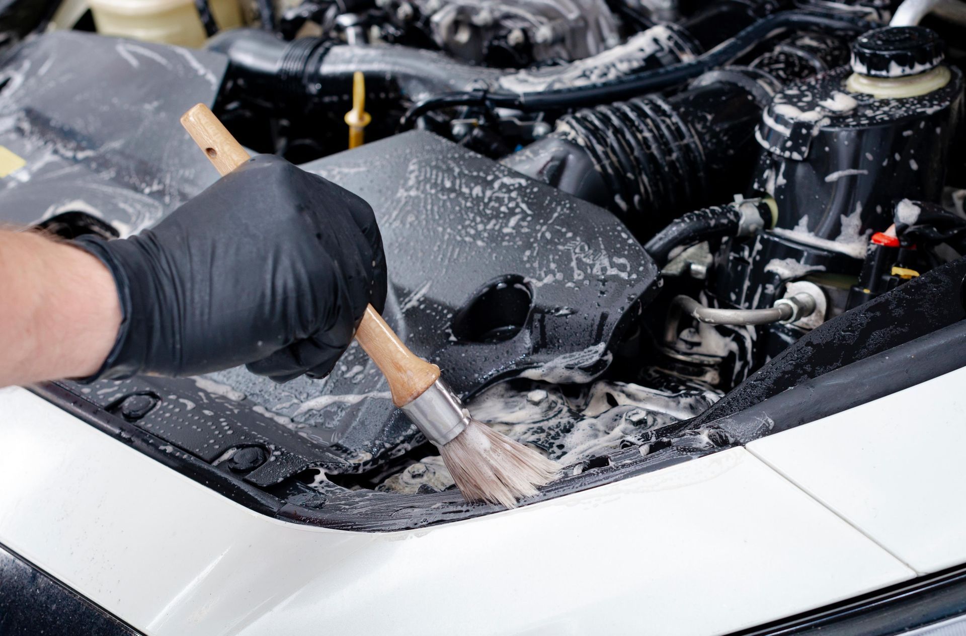 Gloved hand with brush cleaning a car engine bay, covered in soap suds. White car, black engine parts.
