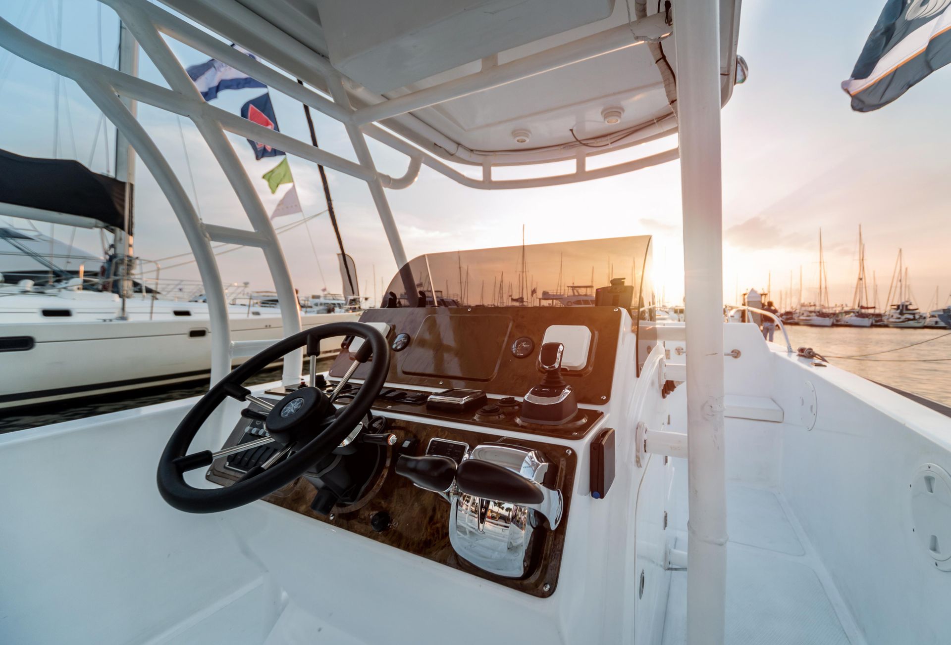 Boat cockpit with steering wheel and controls, overlooking a harbor at sunset.