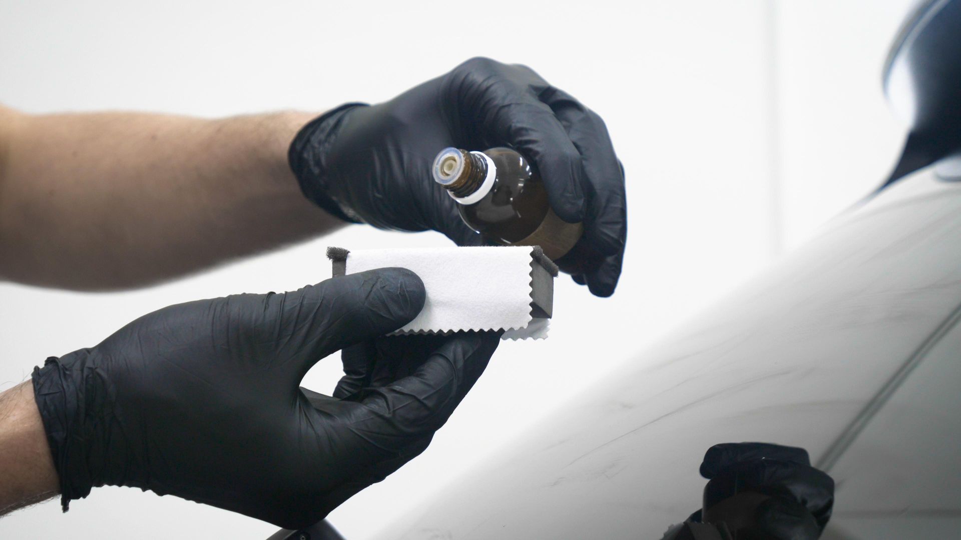 Black-gloved hands applying liquid from a bottle onto a white applicator, near a vehicle.