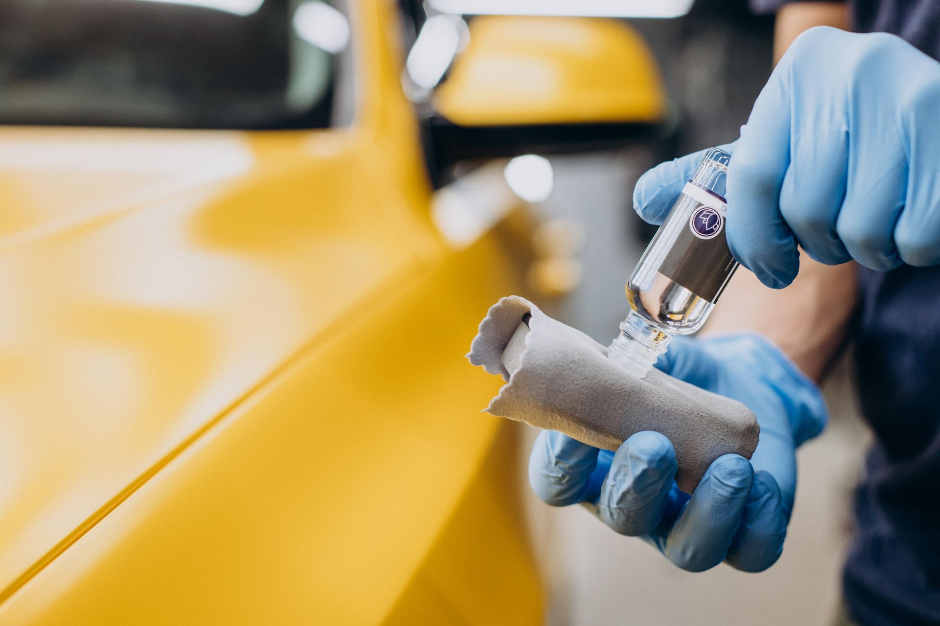 Person applying ceramic coating to a yellow car with a microfiber applicator, wearing blue gloves.