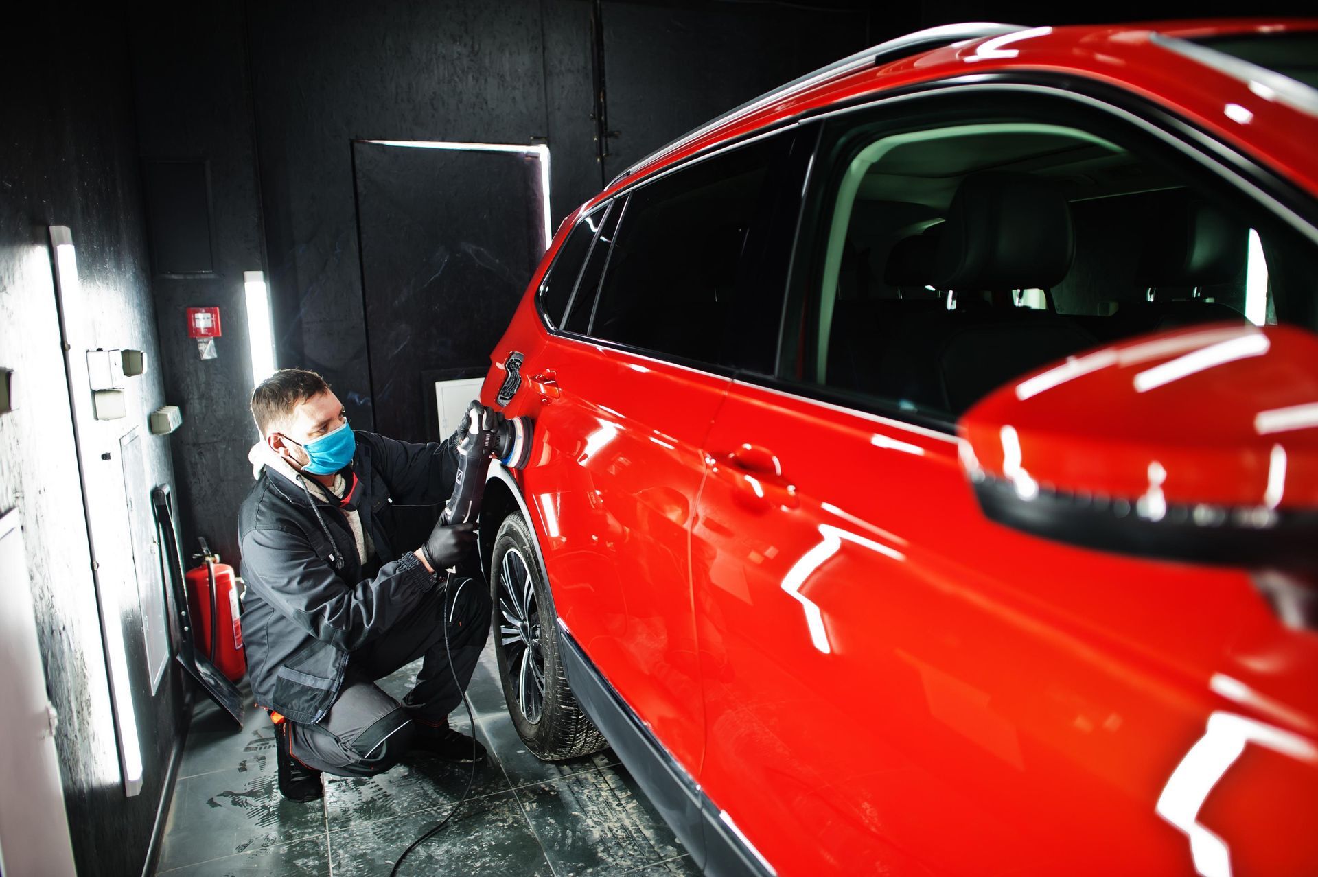 A person in a mask polishing a bright red car inside a workshop.