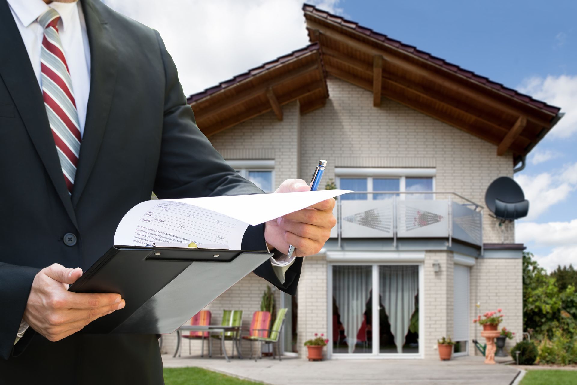 A man in a suit and tie is holding a clipboard in front of a house.