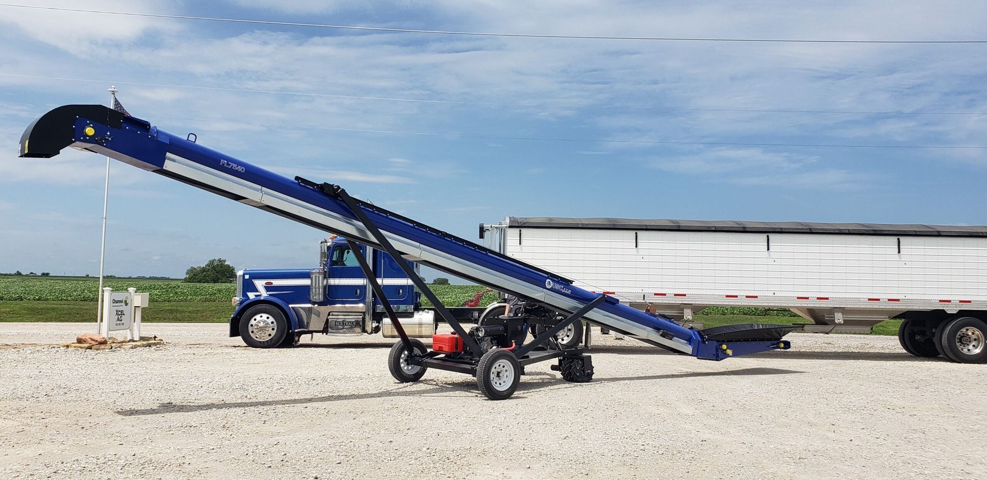 Blue conveyor belt loading grain into a white trailer. A blue truck is parked nearby.