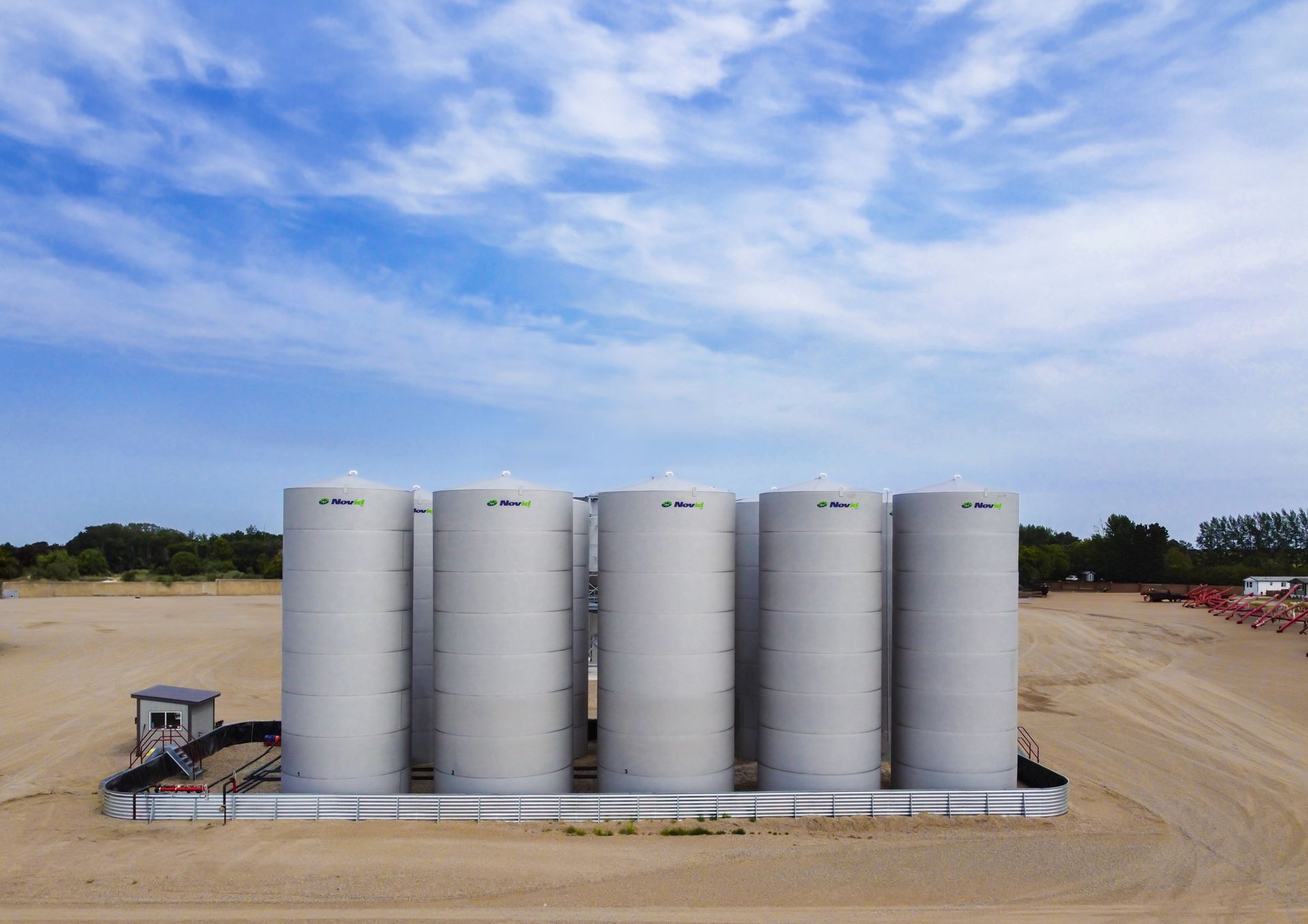 Five large, cylindrical, gray industrial tanks under a blue sky, on a dirt lot.