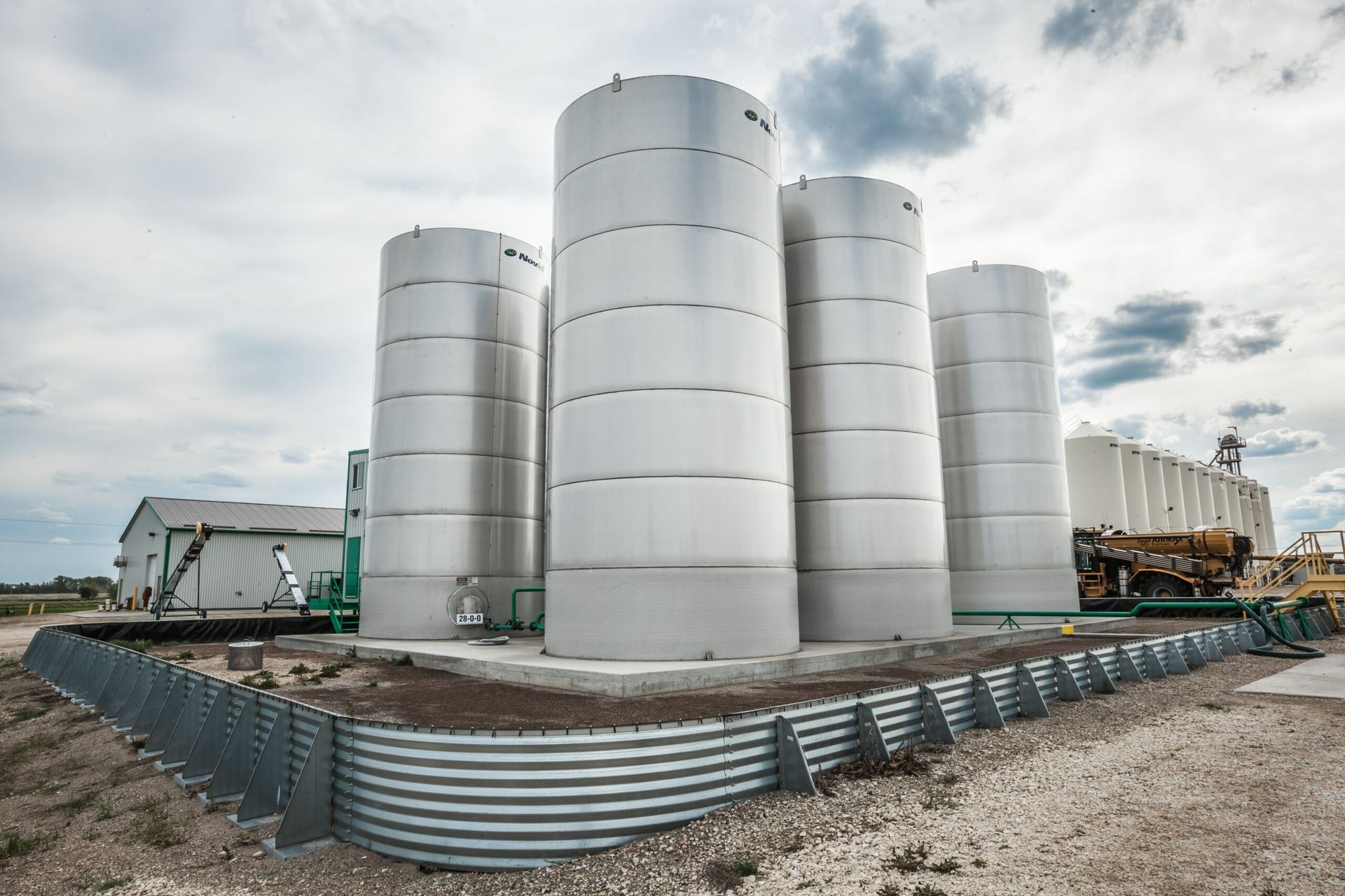 Four large white cylindrical storage tanks surrounded by a metal berm in a rural setting.