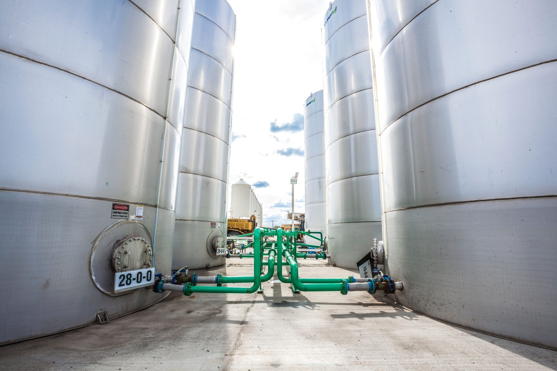 Large, silver industrial tanks with green piping in an outdoor setting under a partly cloudy sky.