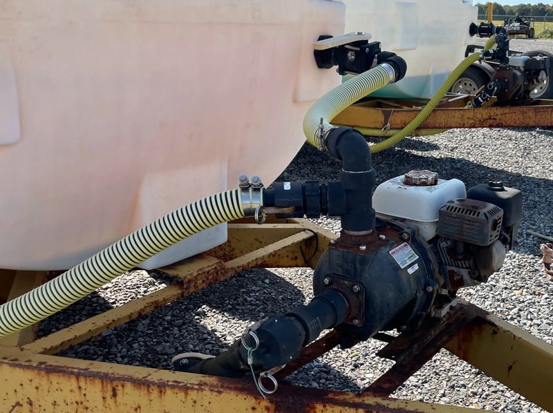 Water tank with attached pump and Honda engine on a yellow trailer in an outdoor setting.