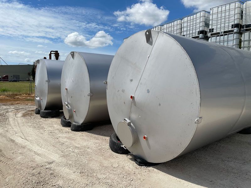 Three large, silver storage tanks on a gravel surface, under a partly cloudy blue sky.