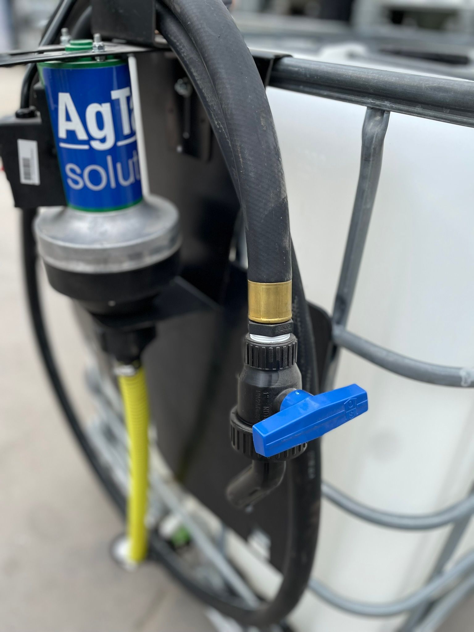 A close-up of a blue-handled valve connected to a black hose, with an AgTrol Solutions pump attached to a white container.