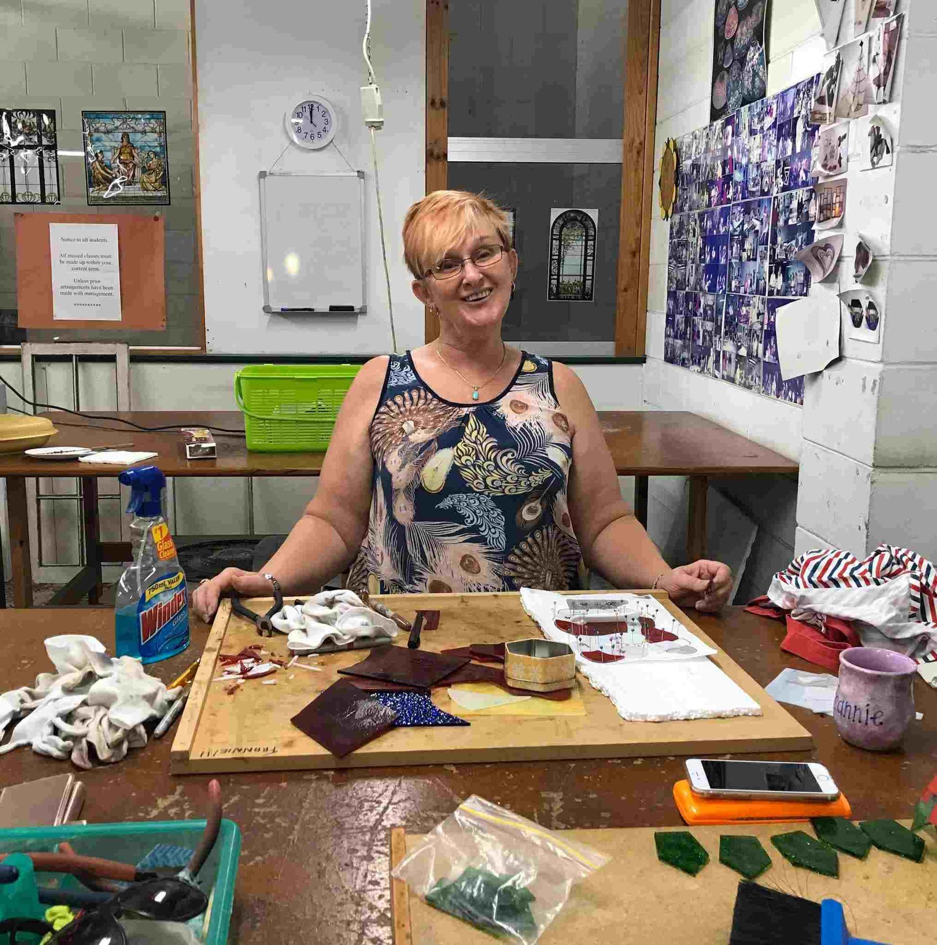 A Woman Is Sitting At A Table In A Room With A Clock On The Wall — Townsville Stained Glass In Mackay, QLD