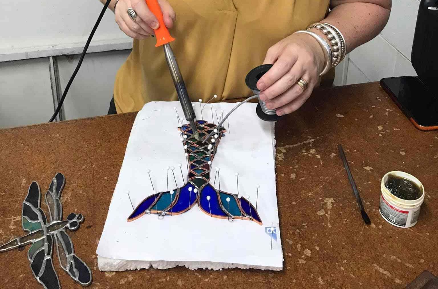 A Woman Is Using A Soldering Iron To Make A Stained Glass Mermaid Tail — Townsville Stained Glass In Charters Towers, QLD