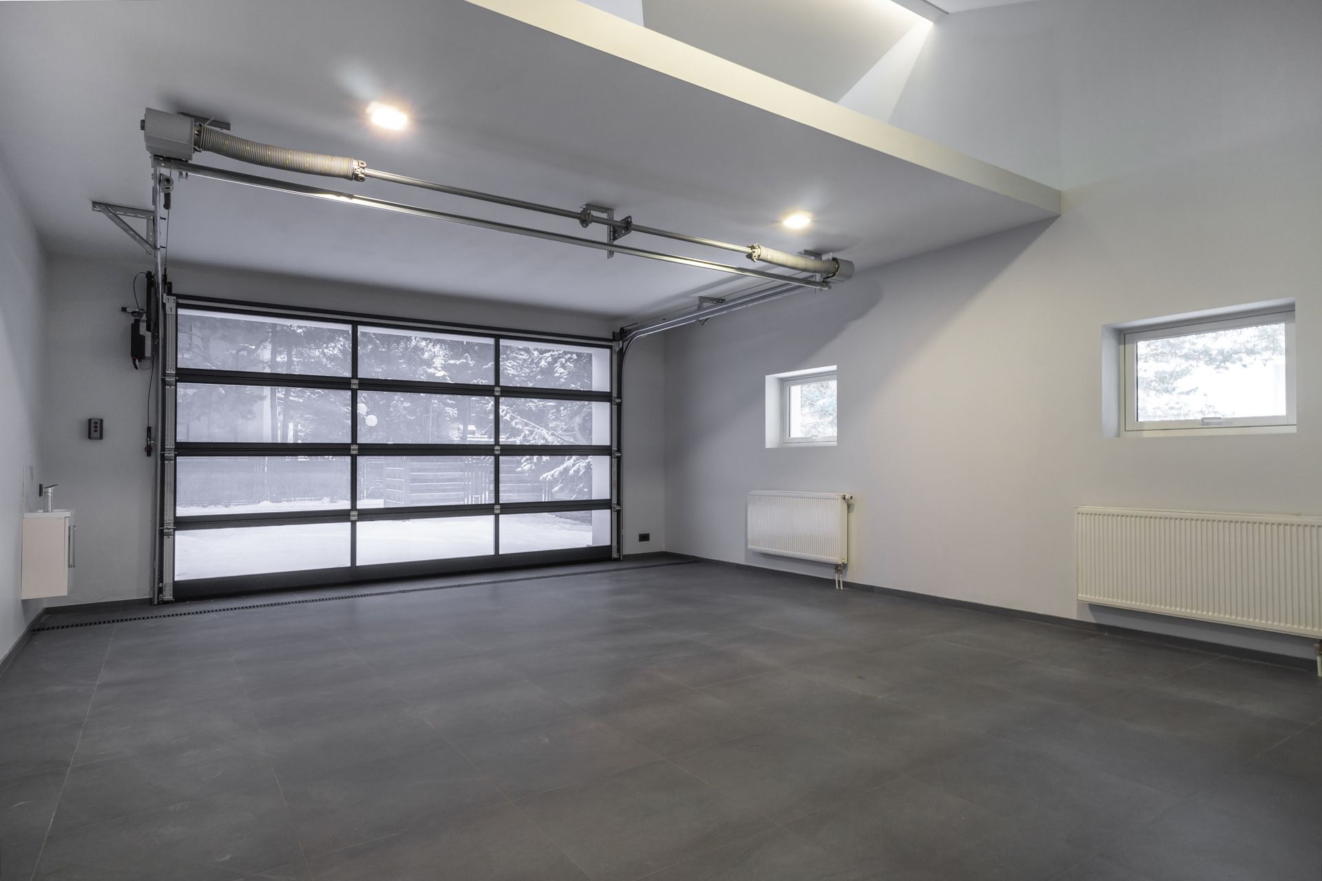 Garage vide et moderne avec porte vitrée et neige visible à l'extérieur. Murs gris, sol noir et deux petites fenêtres.