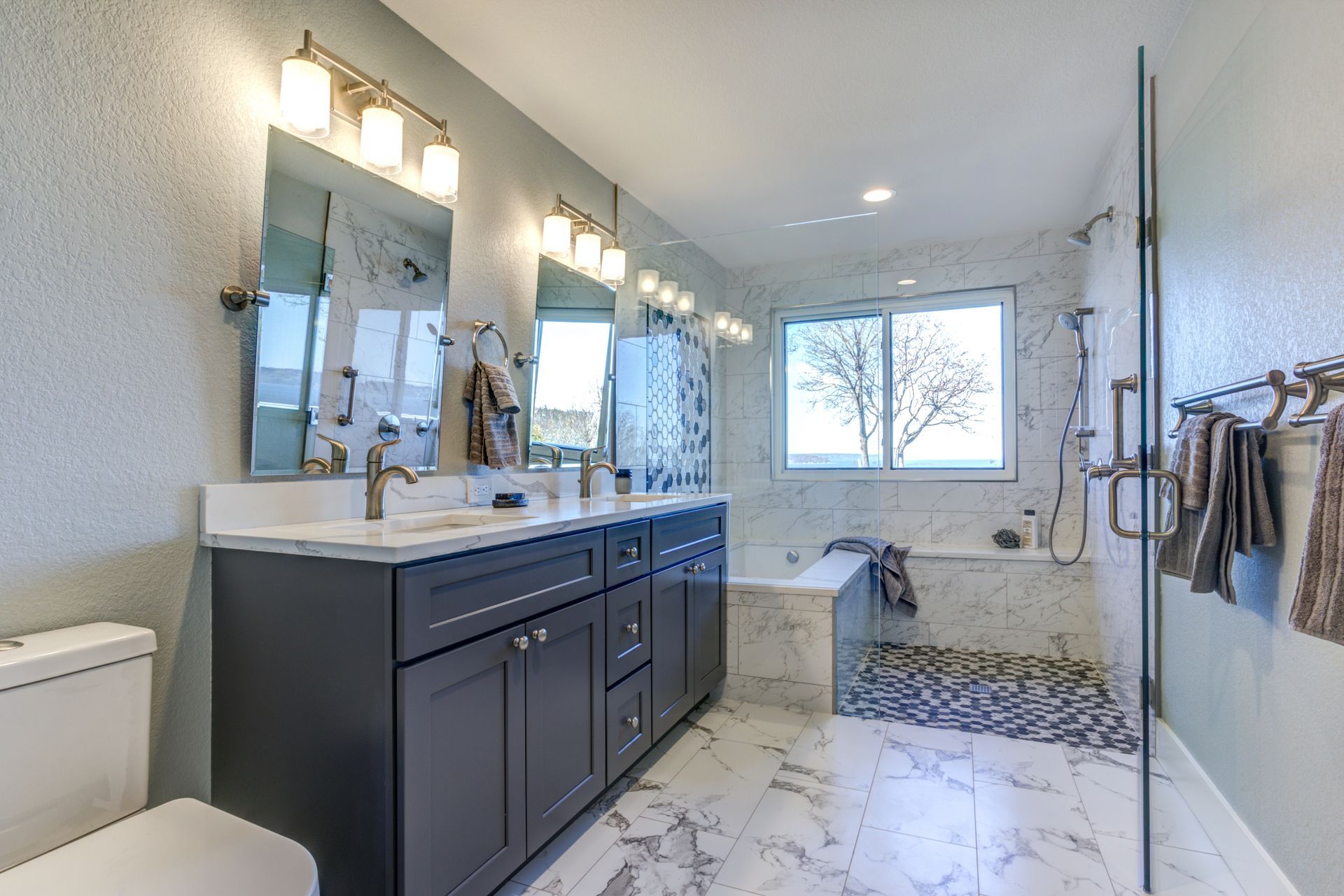 Salle de bain spacieuse et moderne avec vanité grise, sols en marbre et fenêtre donnant sur les arbres.