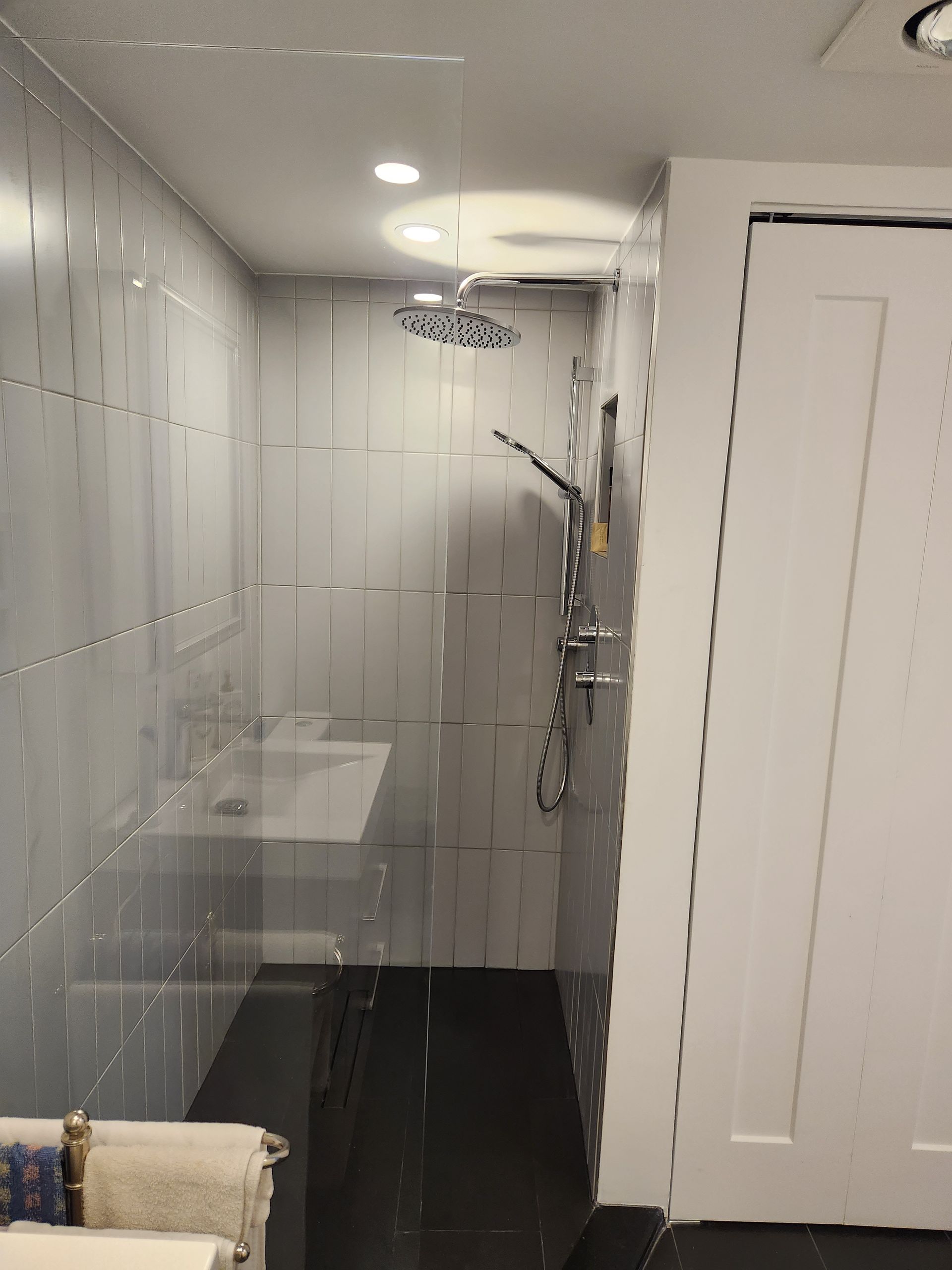 Petite douche carrelée avec porte vitrée, pommeau et robinet. Porte blanche à droite.