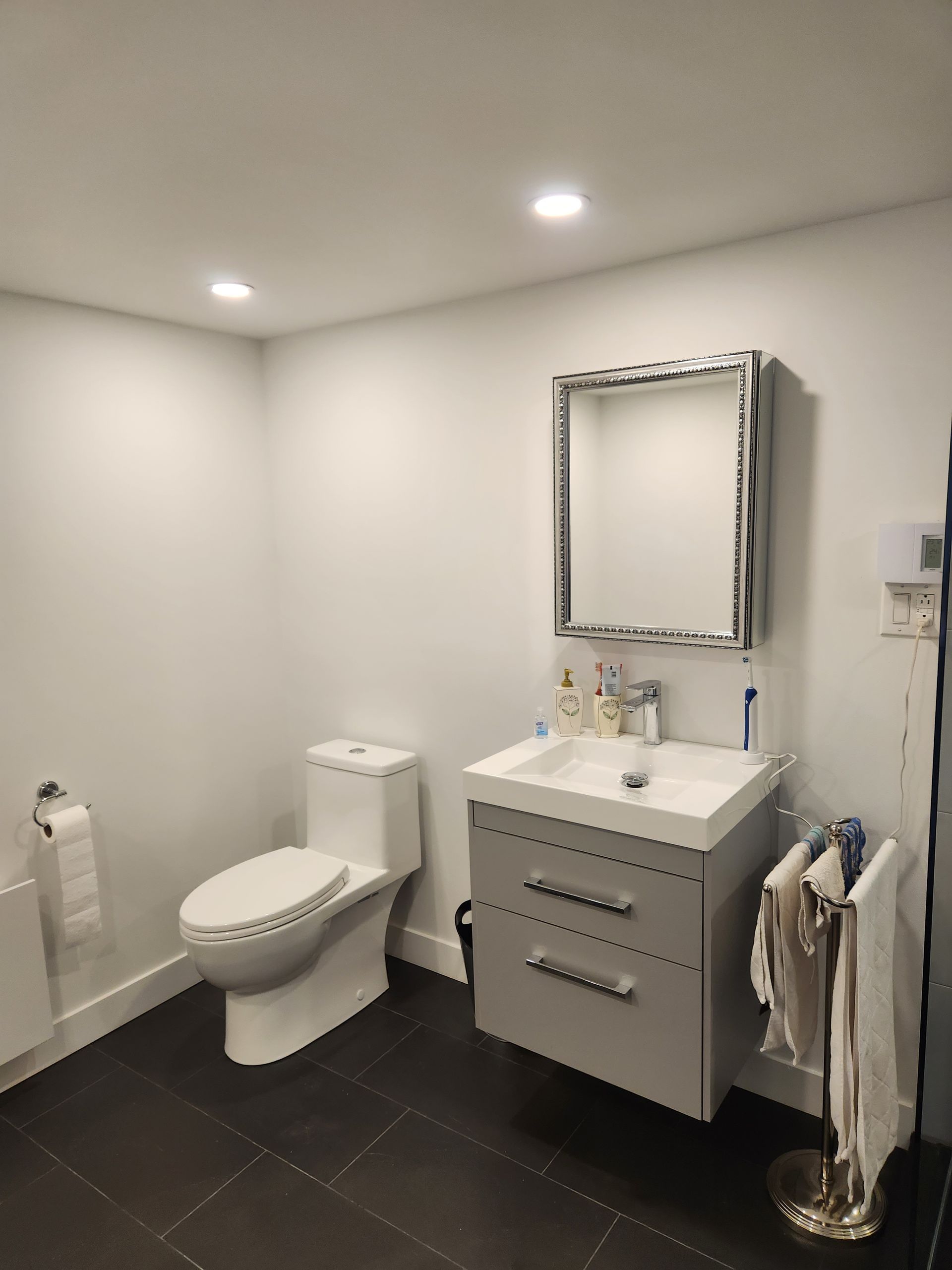 Petite salle de bain blanche avec toilettes, meuble-lavabo, miroir et porte-serviettes. Carrelage foncé au sol.