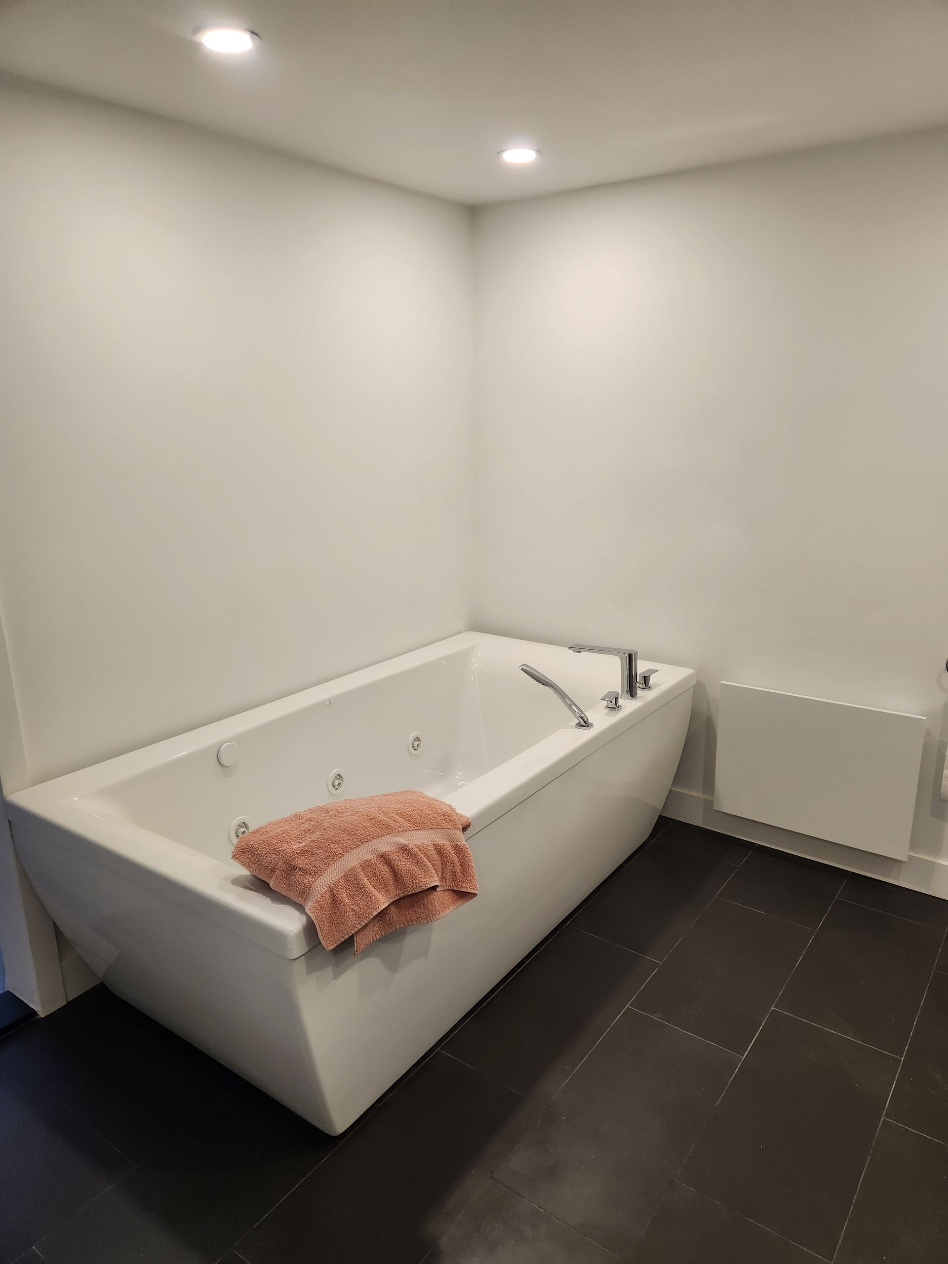 Une baignoire blanche moderne dans un coin, un sol en carrelage noir, des murs blancs et une serviette rose.