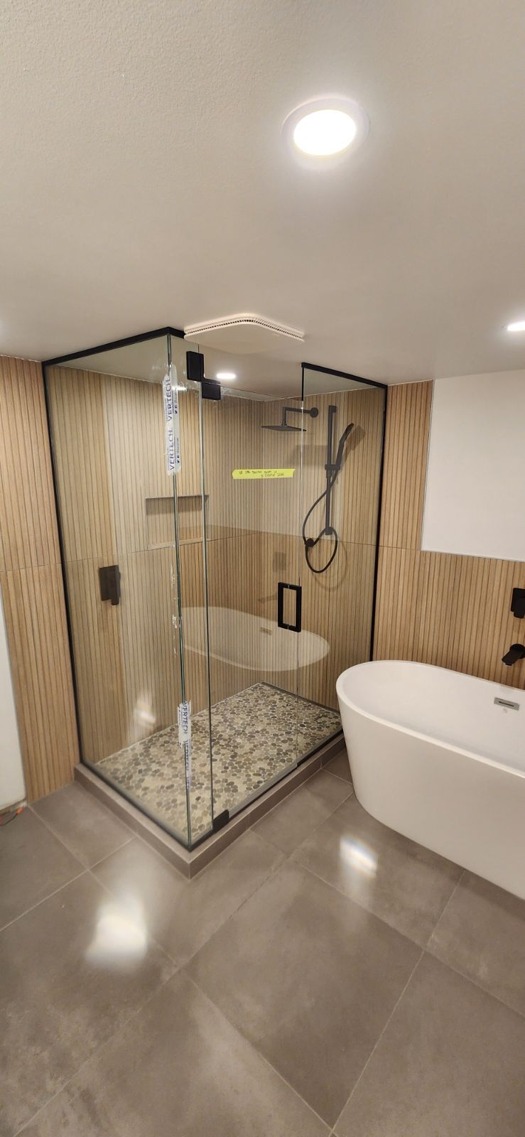 Salle de bain moderne avec douche en verre, baignoire autoportante, sol en carrelage gris, murs aspect bois.