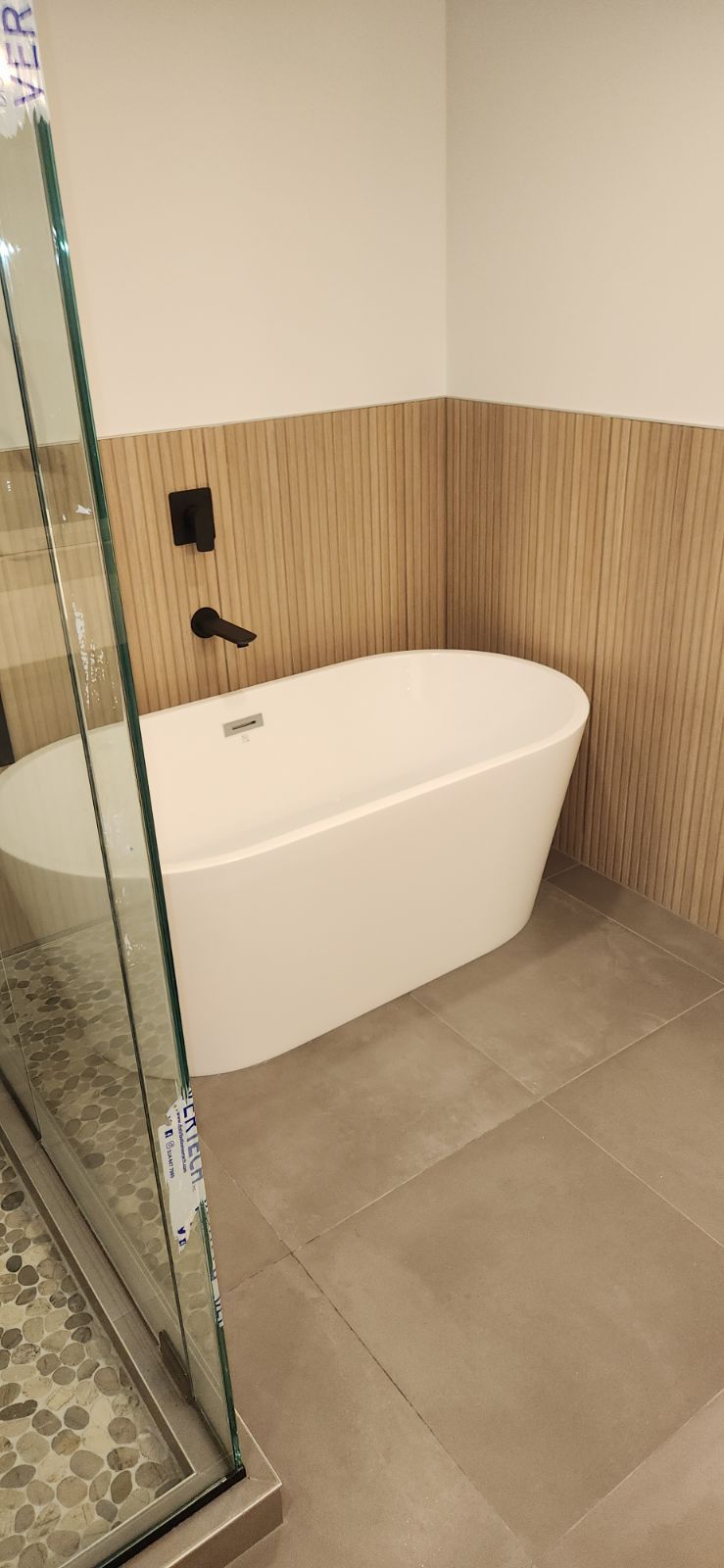 Une baignoire blanche dans une salle de bain avec boiseries et carrelage gris. Une douche en verre se trouve à gauche.