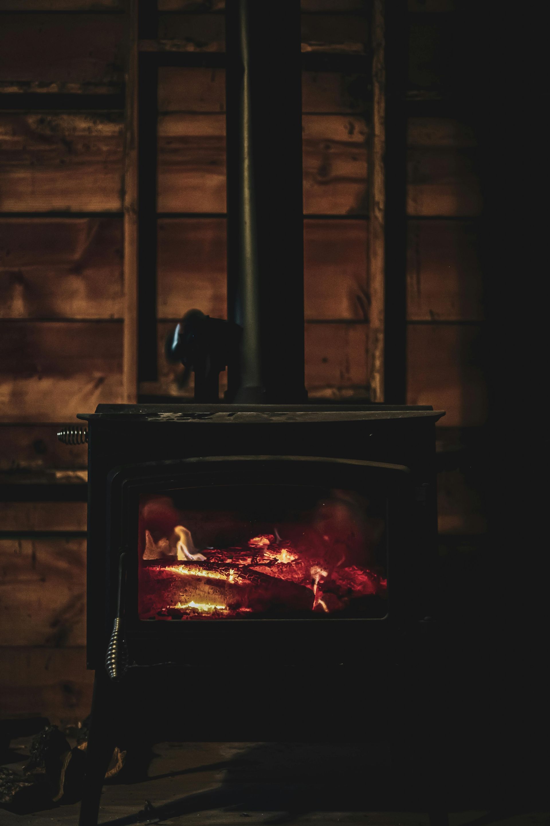 Wood stove burning in a dim rustic cabin, with glowing embers and wooden shelves behind it — Southern Trade Services in Warburton, VIC