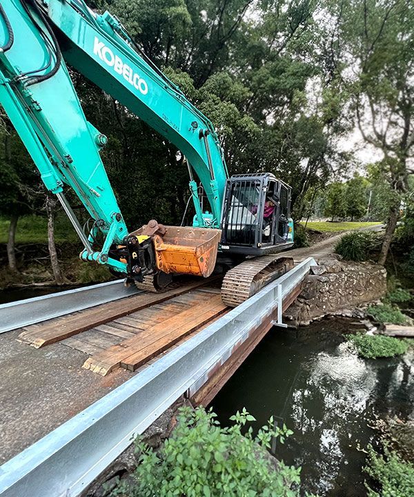 A Green Excavator is Sitting on Top of a Bridge Over a River — Tallow Tree Services Pty Ltd in Bangalow, NSW