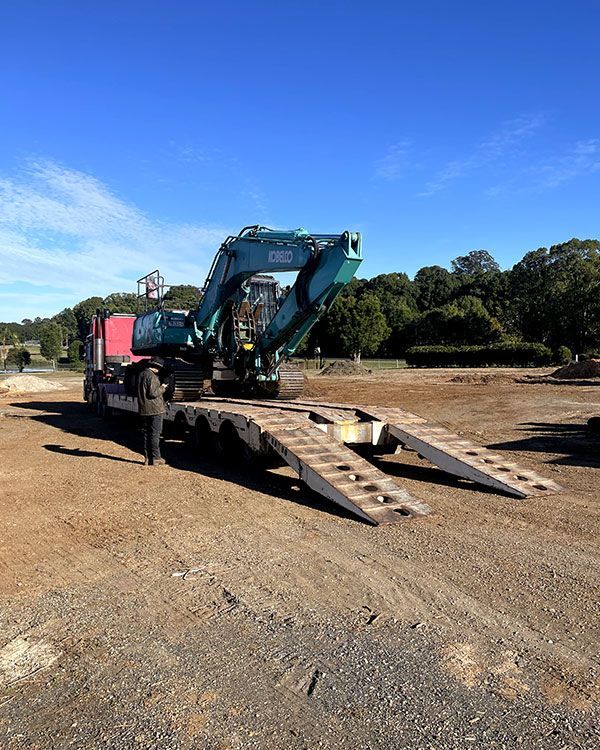 An Excavator is Being Loaded Onto a Trailer in a Dirt Field — Tallow Tree Services Pty Ltd in Bangalow, NSW