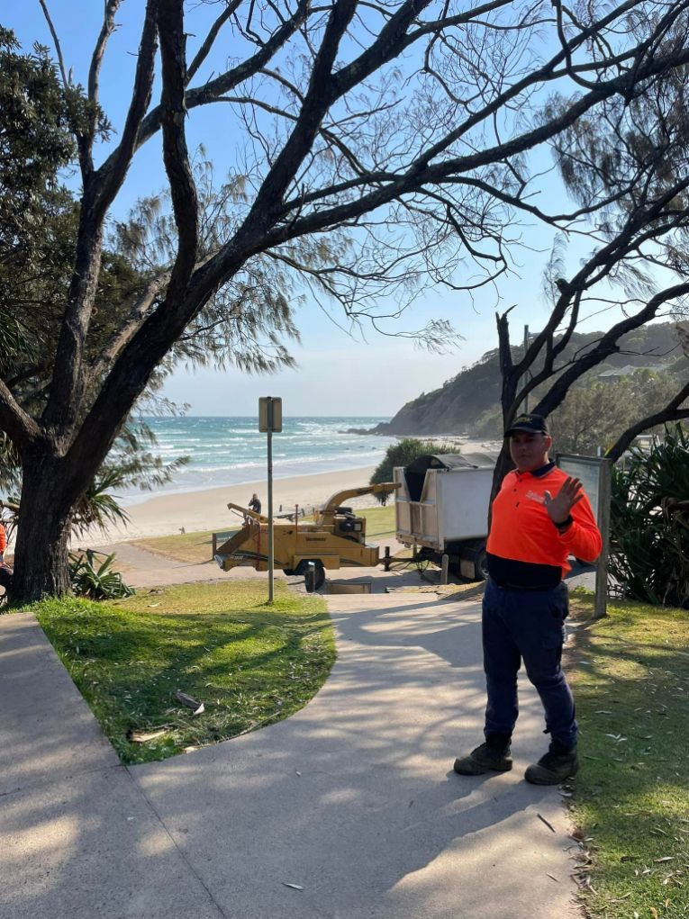 A Man in an Orange Shirt is Standing on a Sidewalk Next to a Beach — Tallow Tree Services Pty Ltd in Casino, NSW