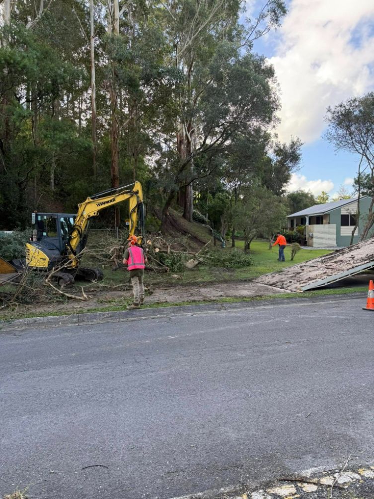 A Man is Standing Next to a Yellow Excavator on the Side of a Road — Tallow Tree Services Pty Ltd in Lennox Head, NSW
