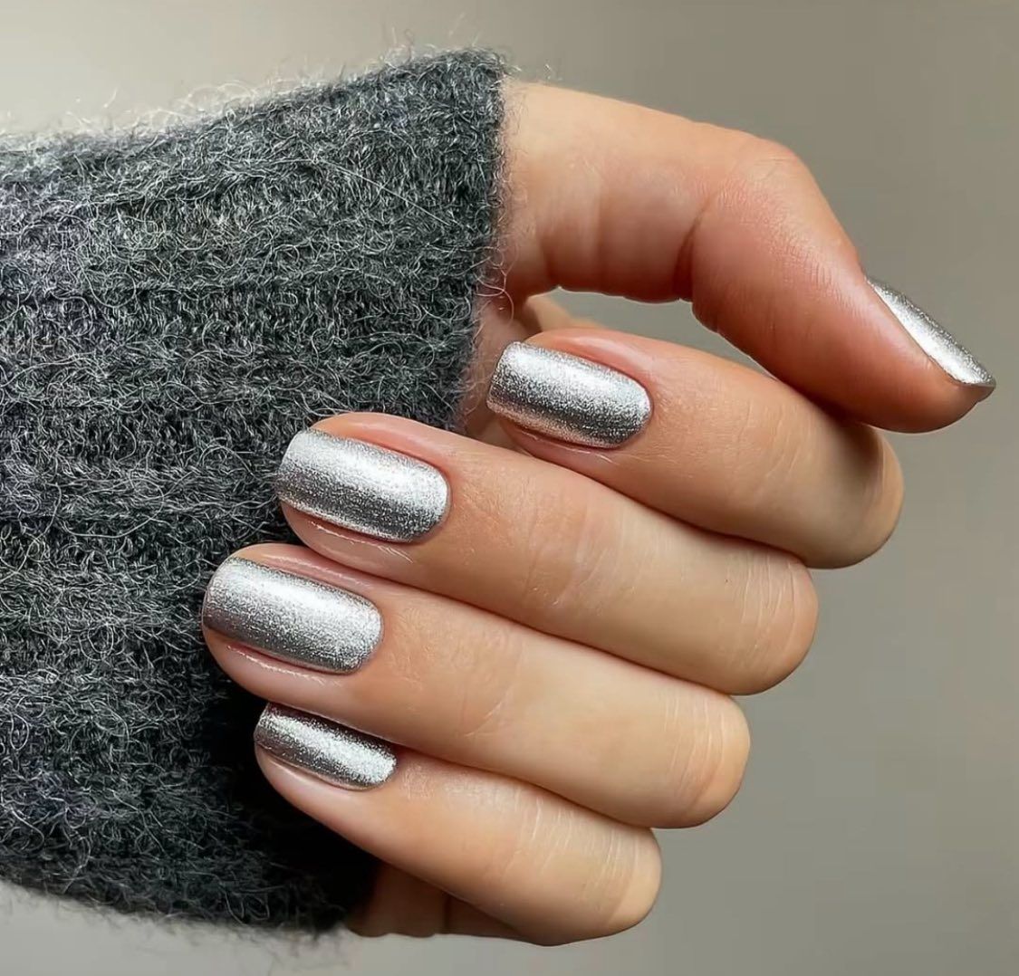 A hand wearing a gray sweater displays short, rounded fingernails painted with a metallic silver, textured polish.