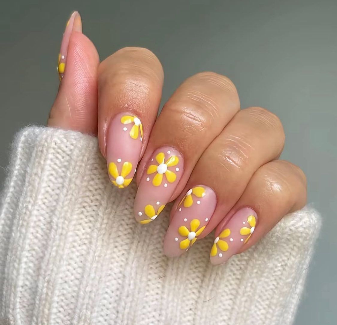 Close-up of almond-shaped nails featuring a light pink base with small, yellow daisy art and white dot accents.