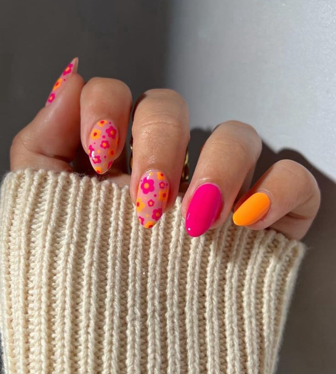 A hand with an almond-shaped manicure featuring vibrant pink and orange floral patterns, solid pink, and solid orange.