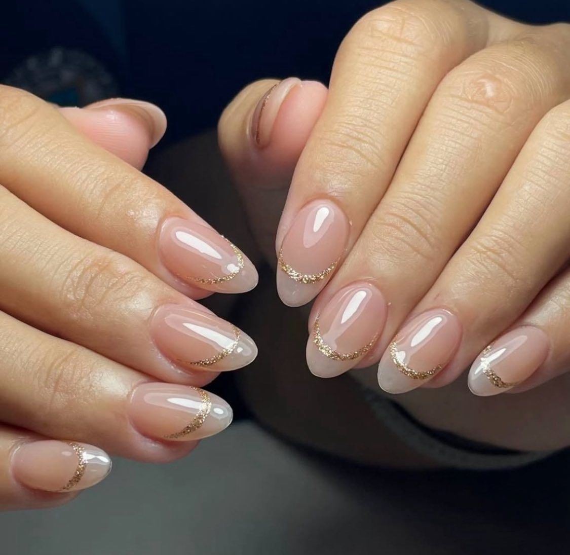 Oval-shaped nails with a sheer nude base and a delicate, sparkling gold glitter crescent at the base of each nail.