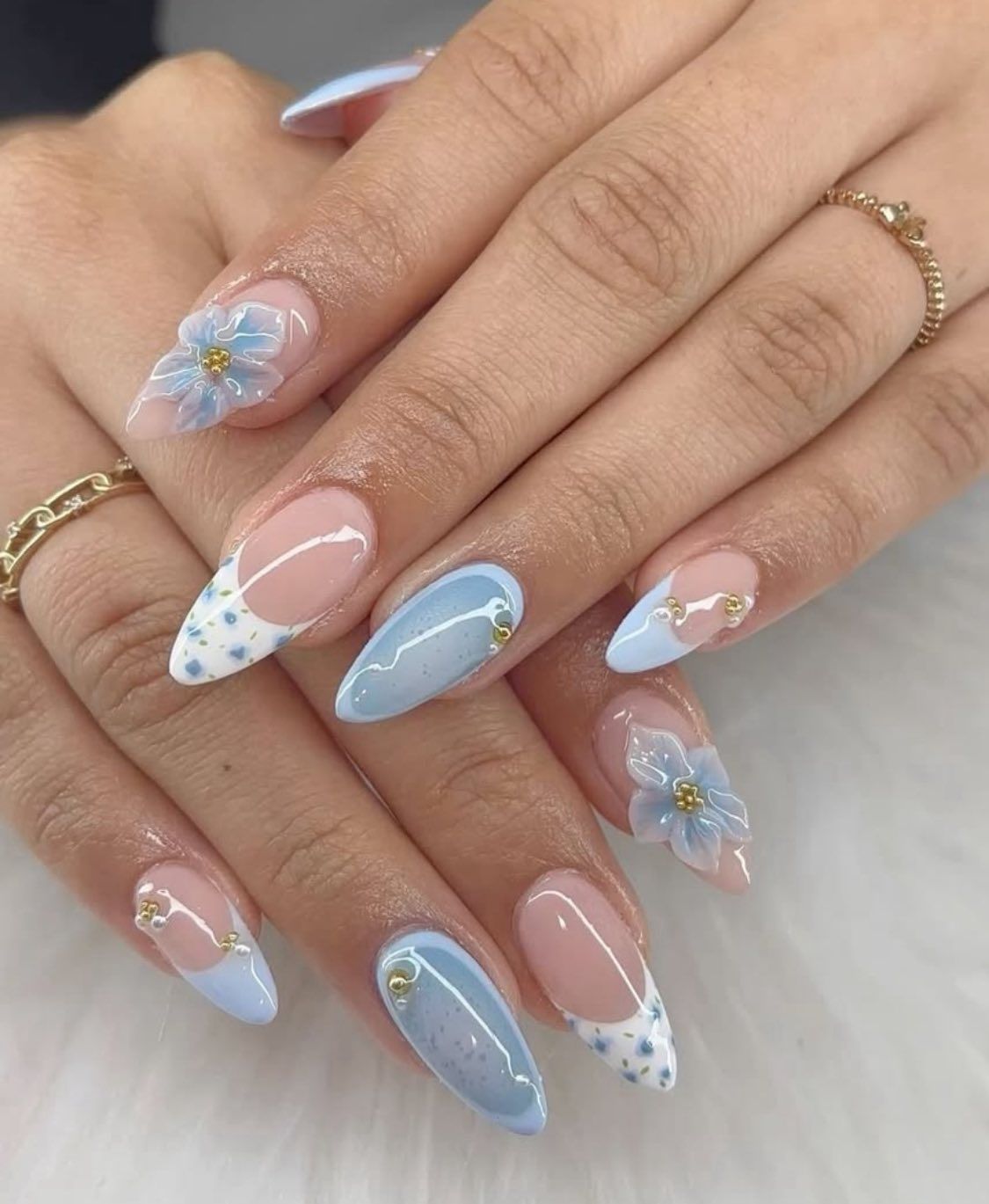 Manicured almond-shaped nails featuring light blue french tips, floral art, and gold accents on a soft nude base.