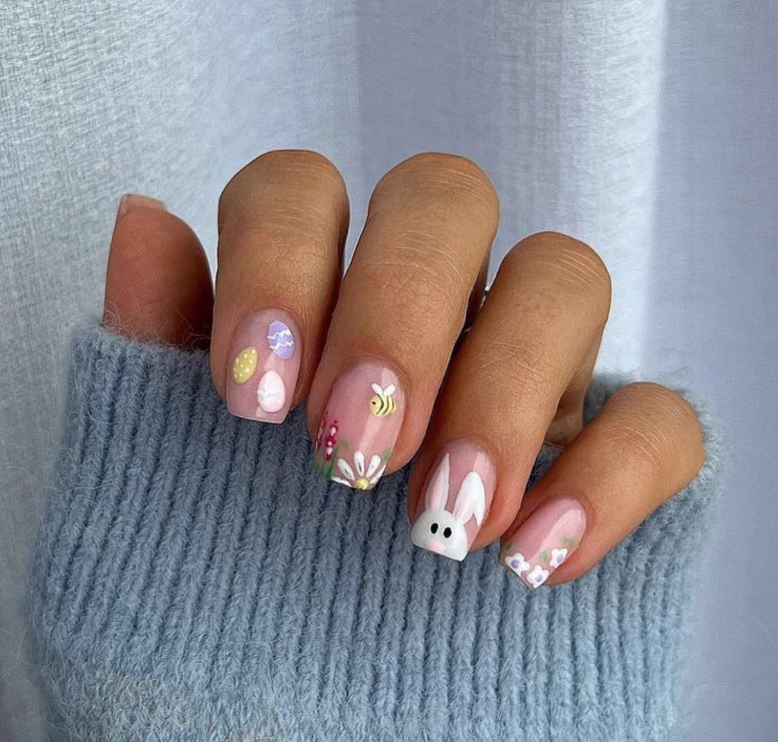 A hand wearing a blue sweater features Easter-themed nail art with pastel eggs, a bee, a daisy, a bunny, and small flowers.
