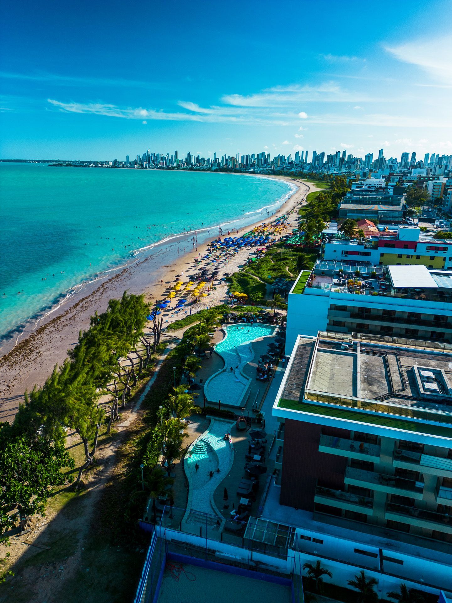 Vista aérea de uma praia com água turquesa, ladeada por edifícios. Um hotel com piscina está em primeiro plano.