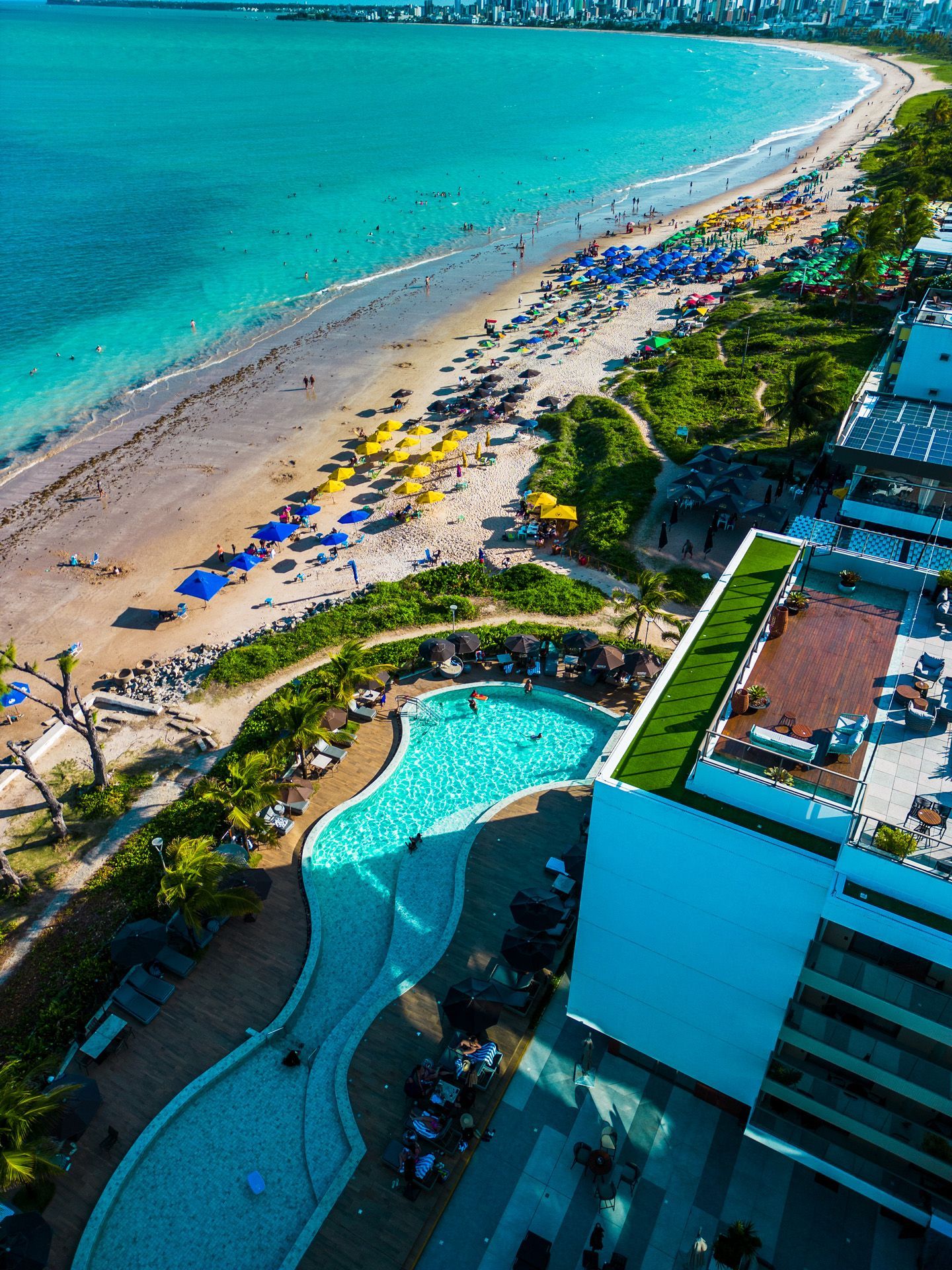 Vista aérea de uma praia com água turquesa, faixa de areia e uma piscina curva ao lado de um edifício moderno.