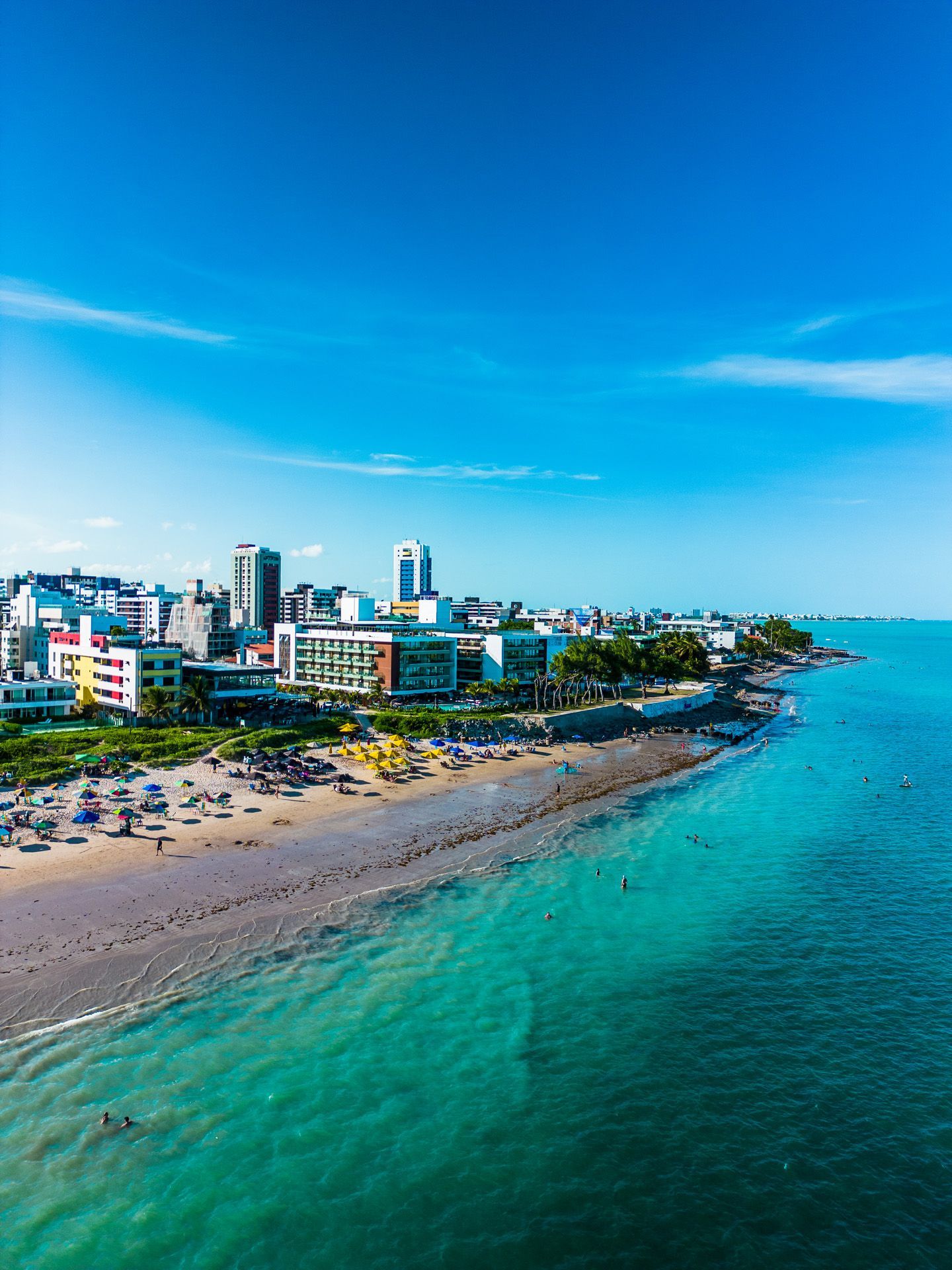 Vista de uma cidade costeira com praia, água turquesa, edifícios e céu azul.