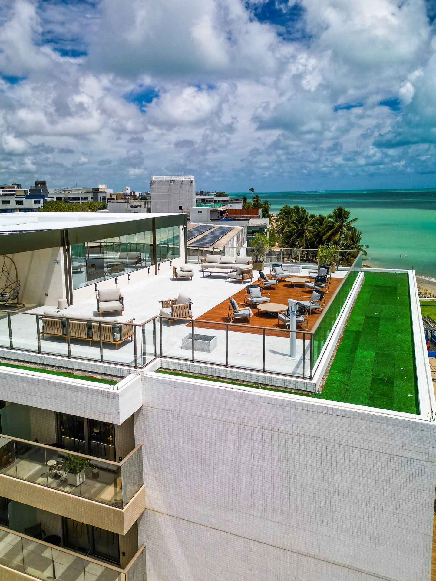 Lounge na cobertura com mesas, cadeiras e grama artificial, com vista para o oceano sob um céu nublado.