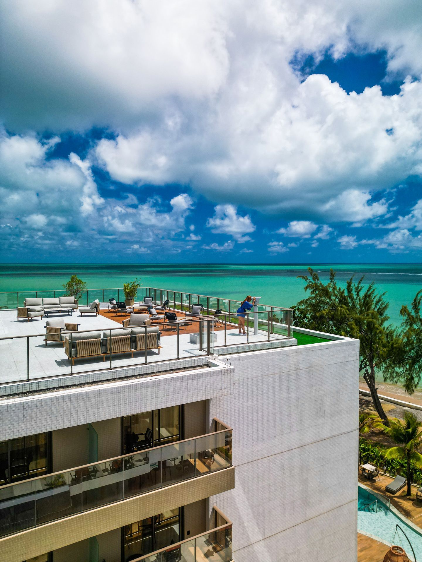 Lounge na cobertura com vista para o mar: prédio branco, água azul-turquesa, céu nublado, mesas ao ar livre.