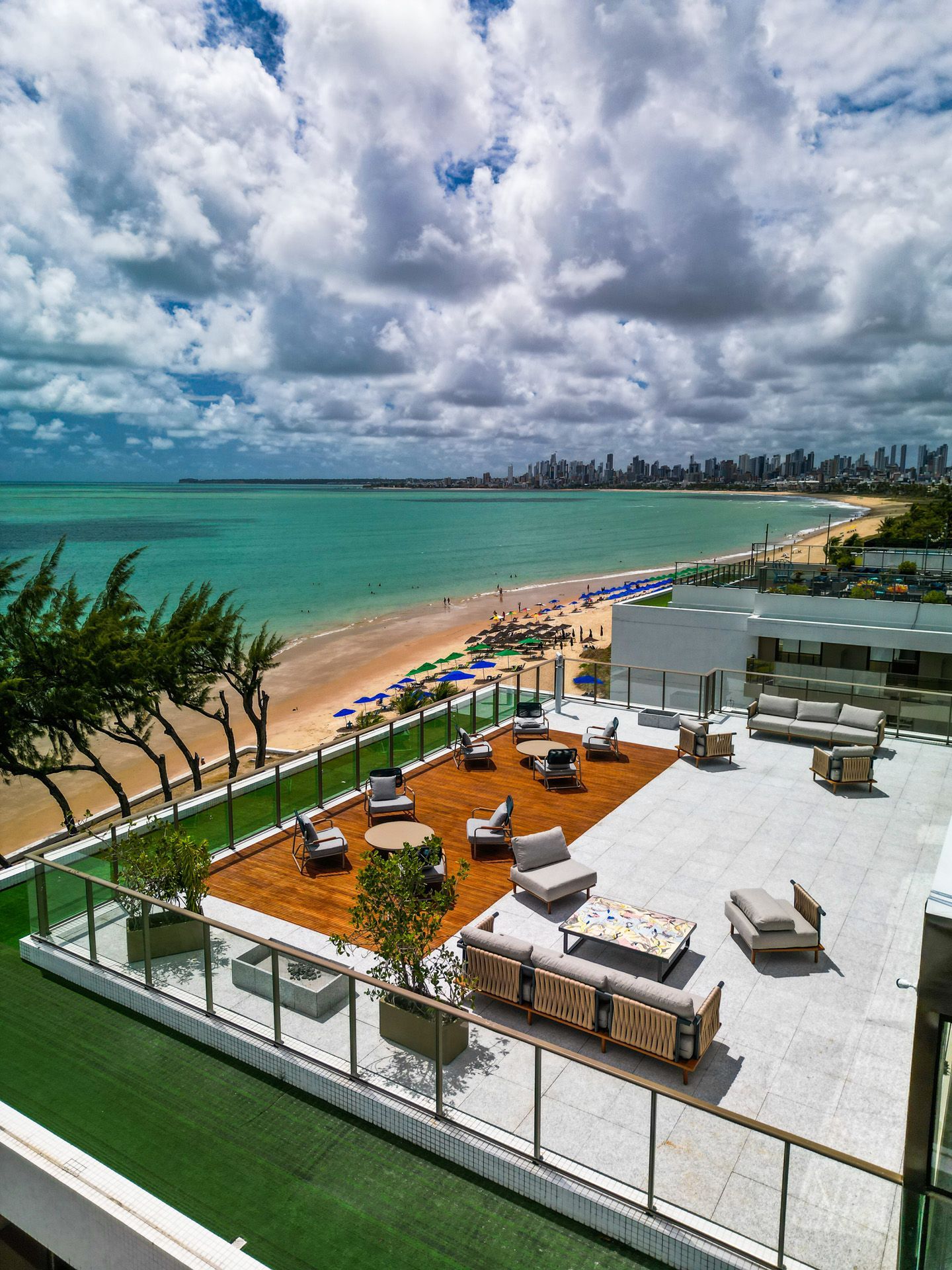 Lounge na cobertura com vista para uma praia e oceano de águas turquesa, com assentos e uma paisagem urbana ao longe sob um céu nublado.