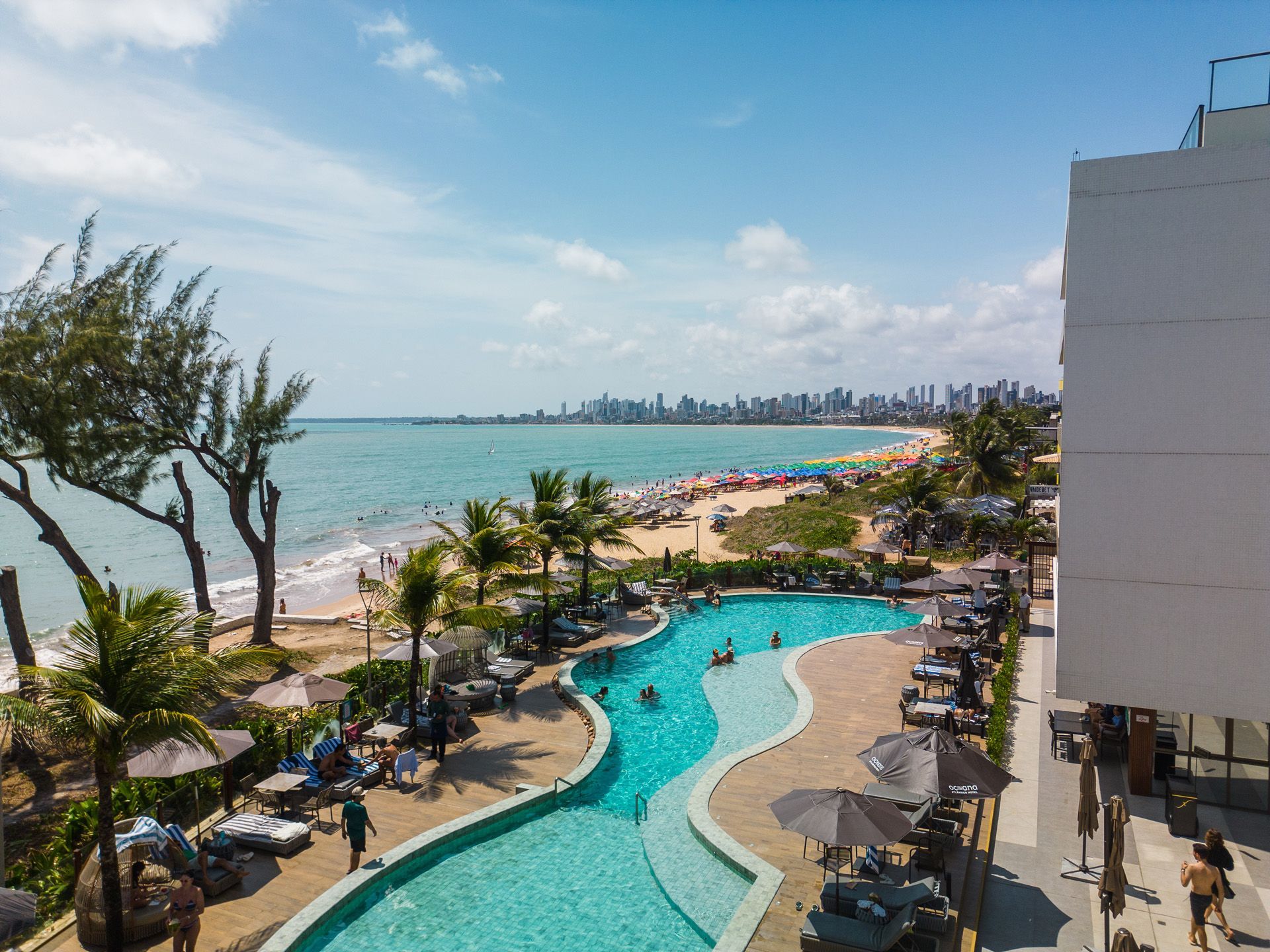 Piscina do hotel com vista para uma praia com pessoas, palmeiras e o horizonte da cidade sob um céu ensolarado.