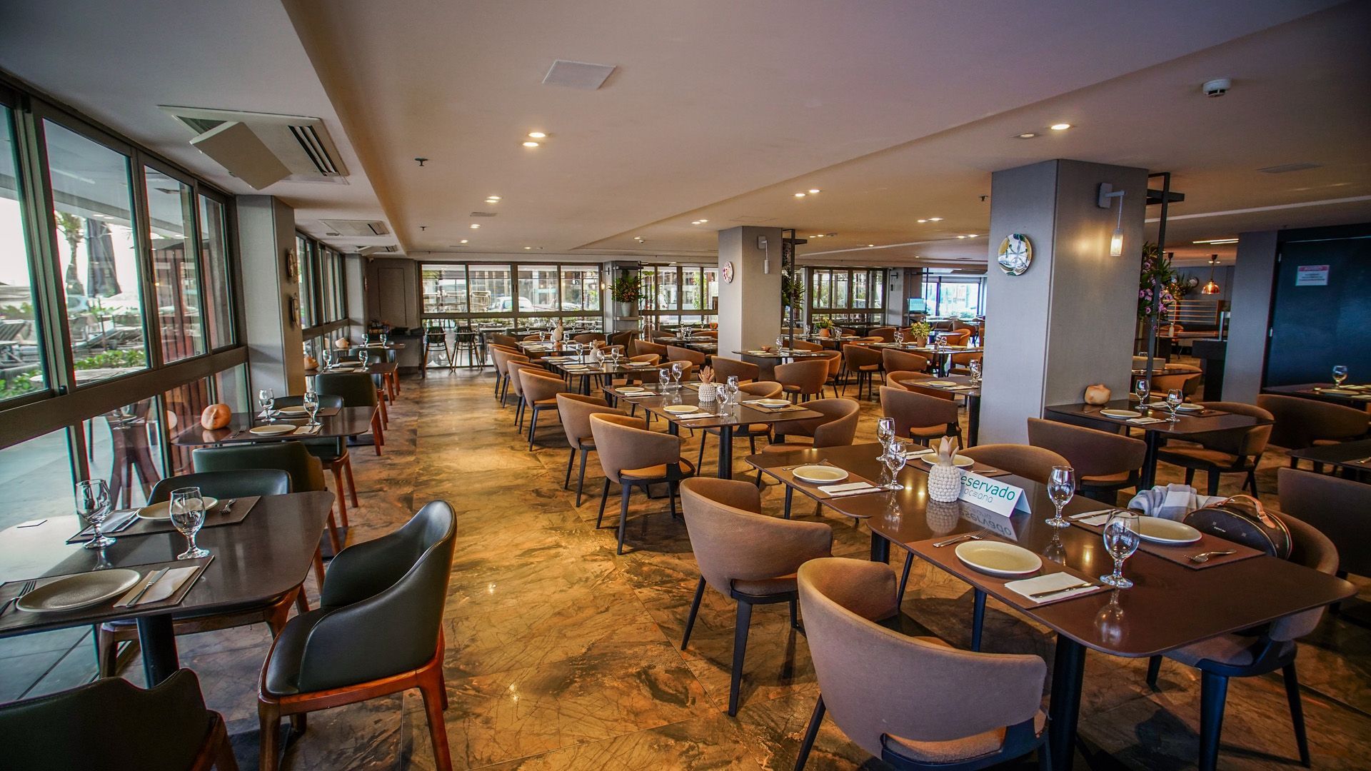 Interior de restaurante com mesas e cadeiras perto de grandes janelas com vista para as árvores ao pôr do sol.